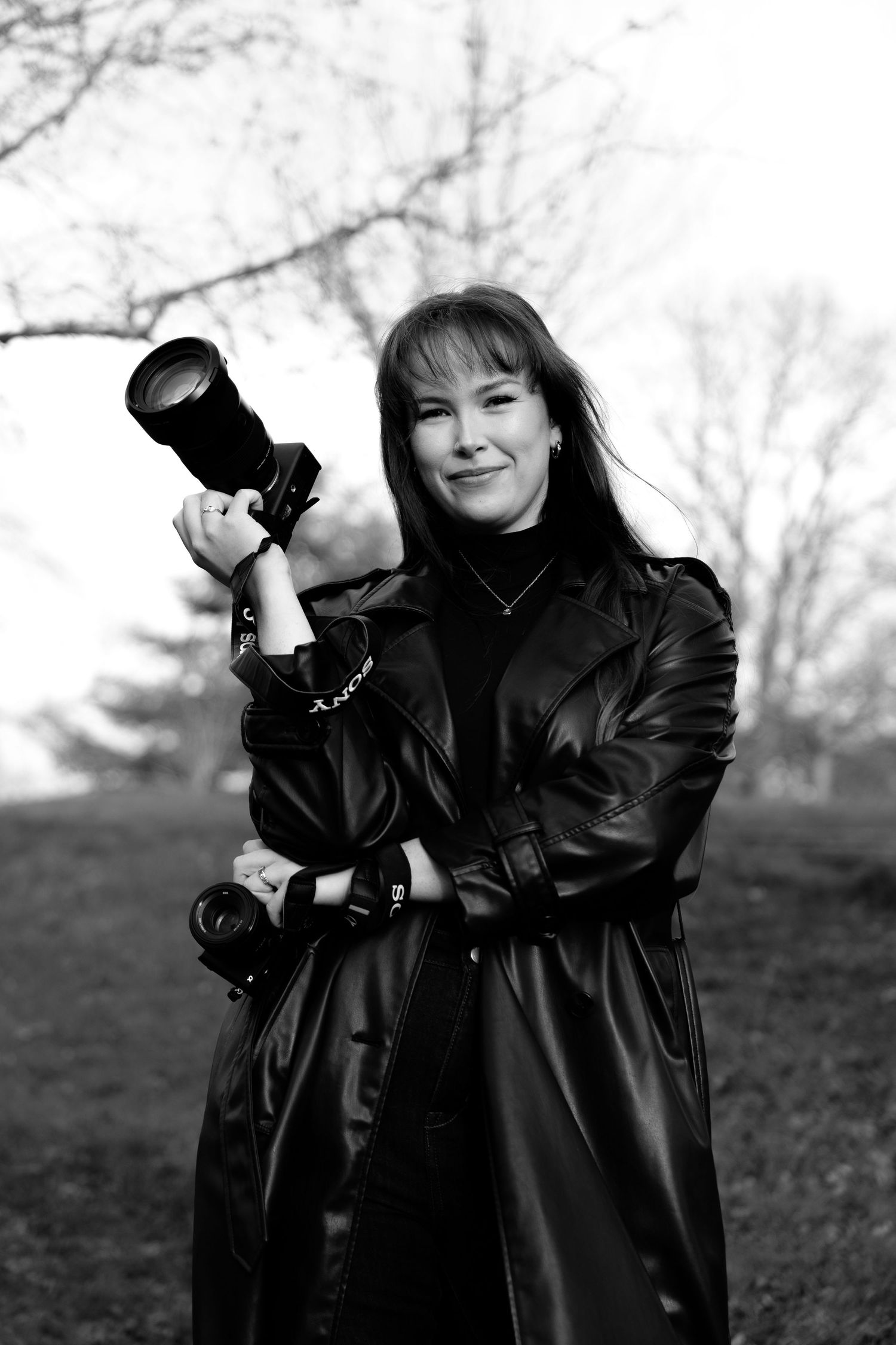 Black and white portrait of a smiling woman in leather coat holding a camera outdoors.