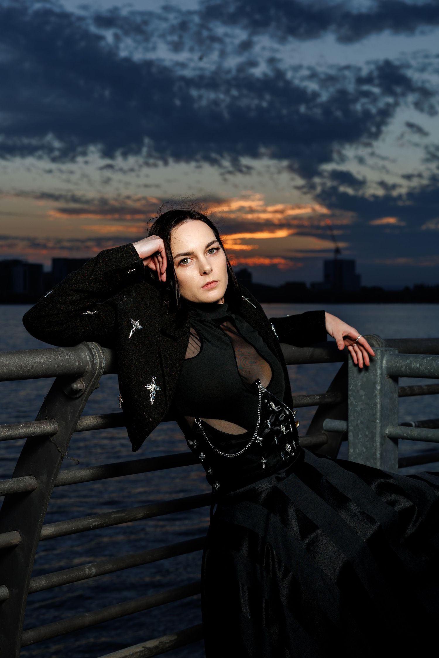Woman in black gothic outfit poses against metal railing at sunset over water.
