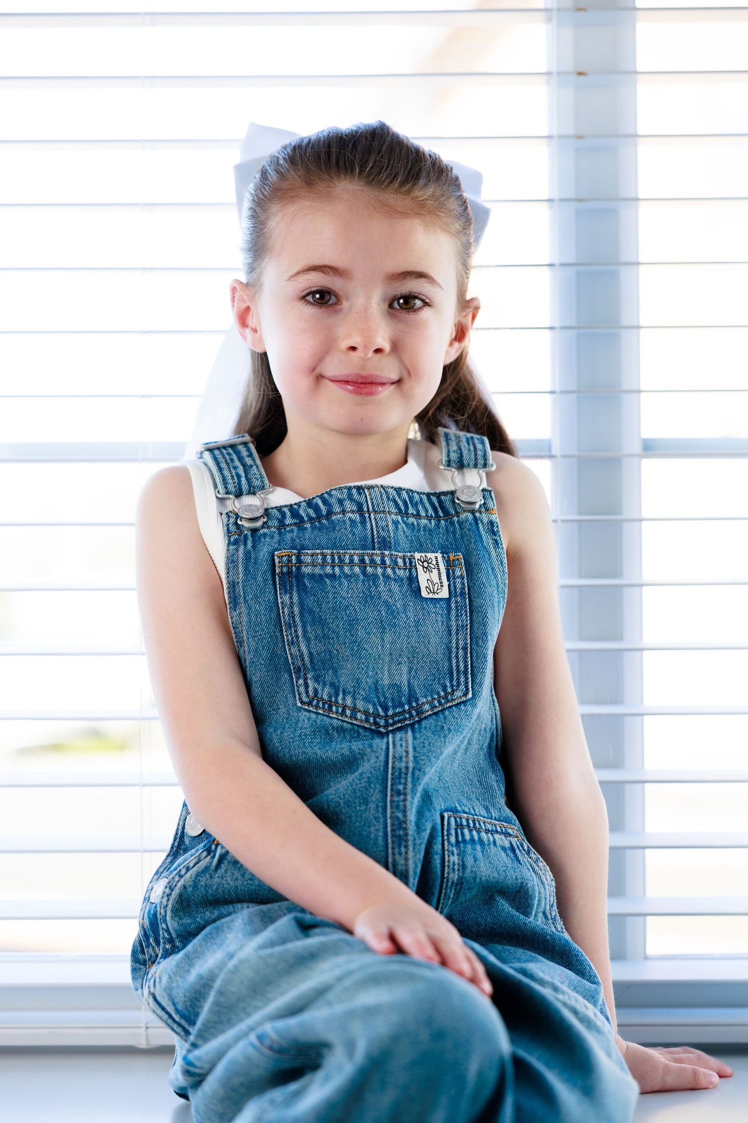 Young girl in denim overalls with white bow sits smiling by window blinds.