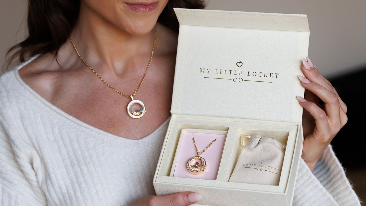 Woman holding an open gift box displaying a My Little Locket Co. gold locket necklace in elegant cream packaging.