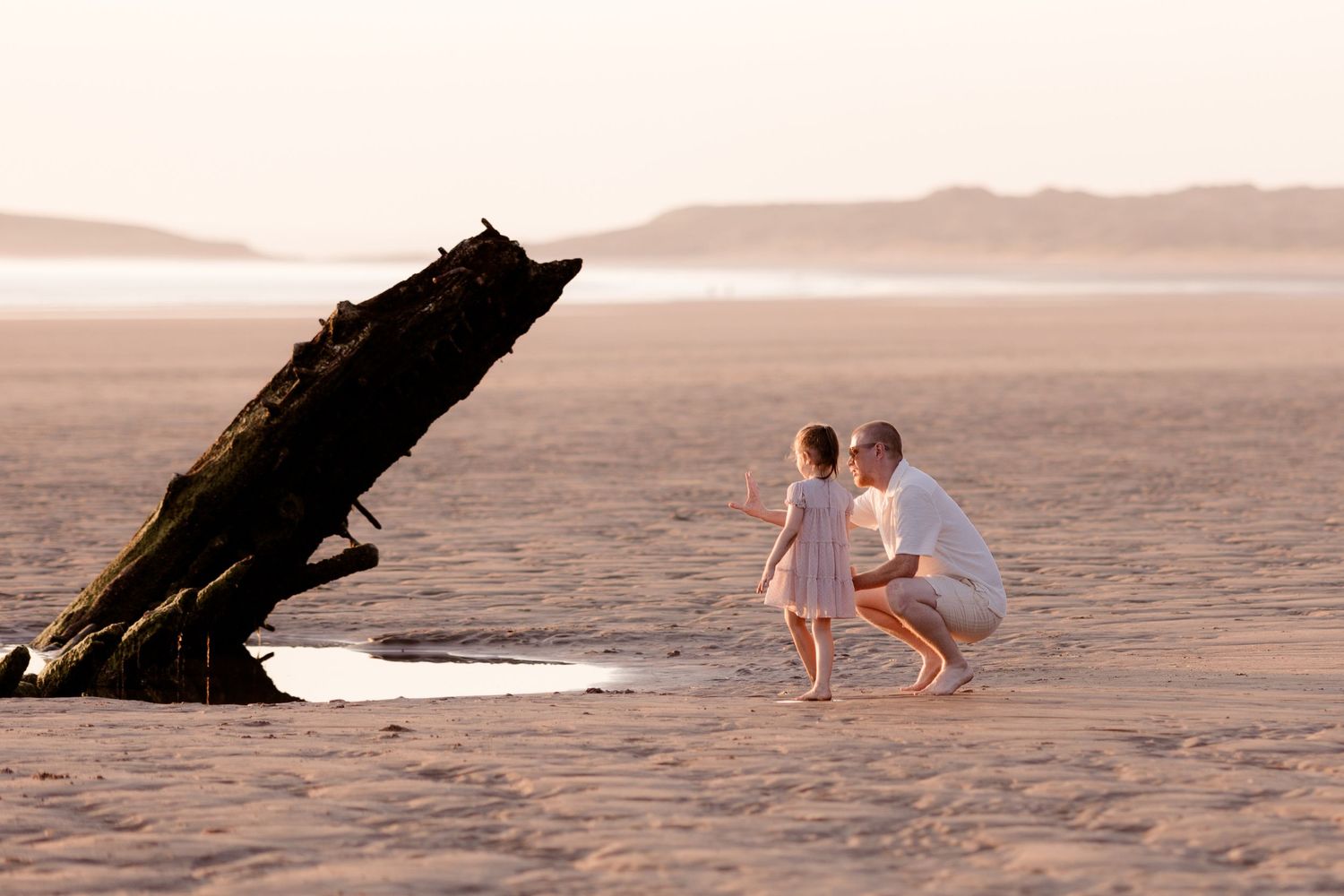 Father and child crouching on a sandy beach near a shipwreck at sunset.