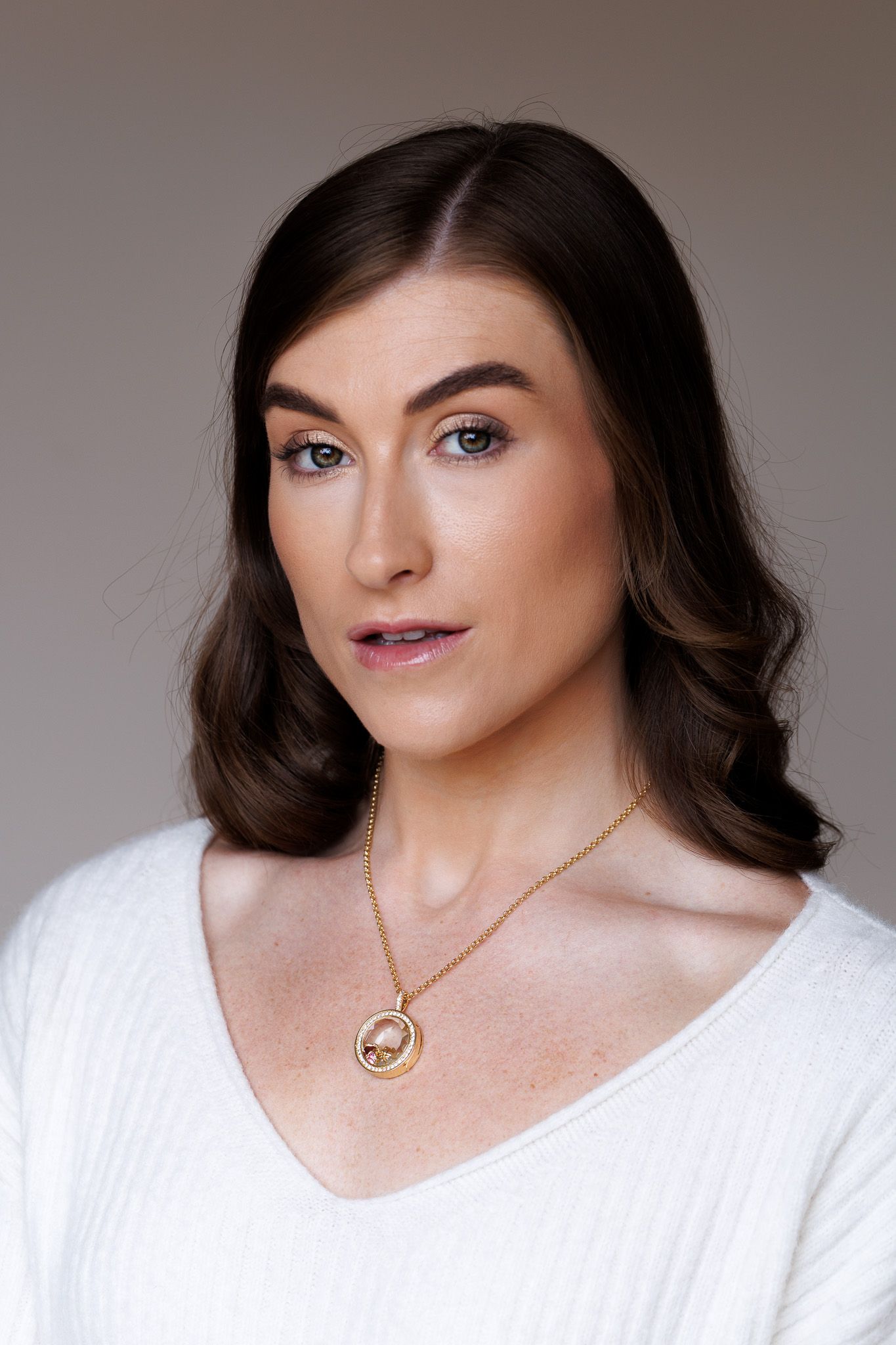 Woman with dark hair wearing a white sweater and gold pendant necklace poses for a portrait photo.