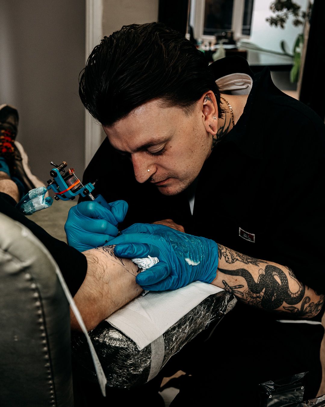 Tattoo artist with sleeve tattoos applying ink to client using blue gloves and tattoo machine.