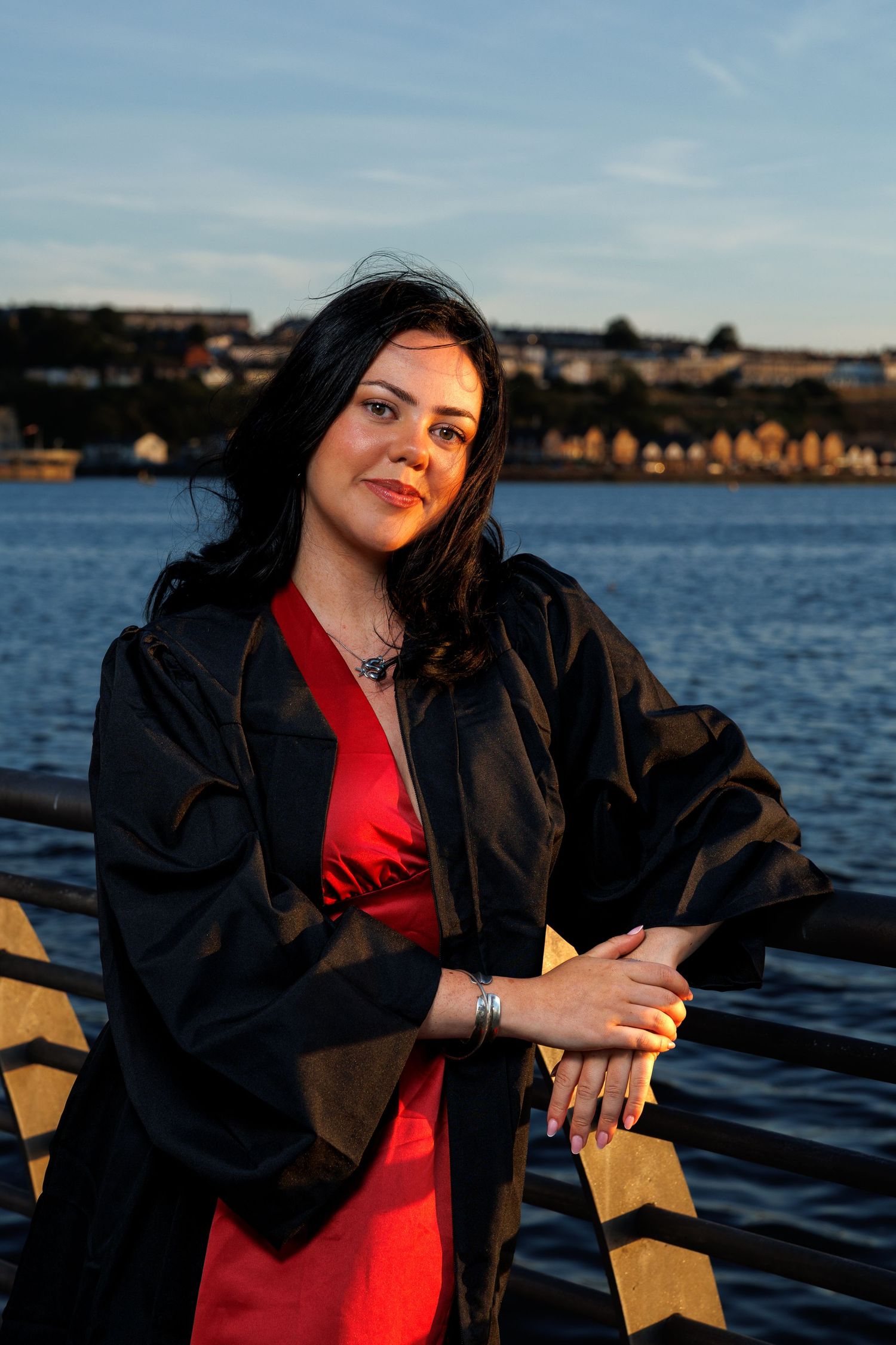 Woman in red dress and black graduation gown leaning on railing by waterfront at sunset.