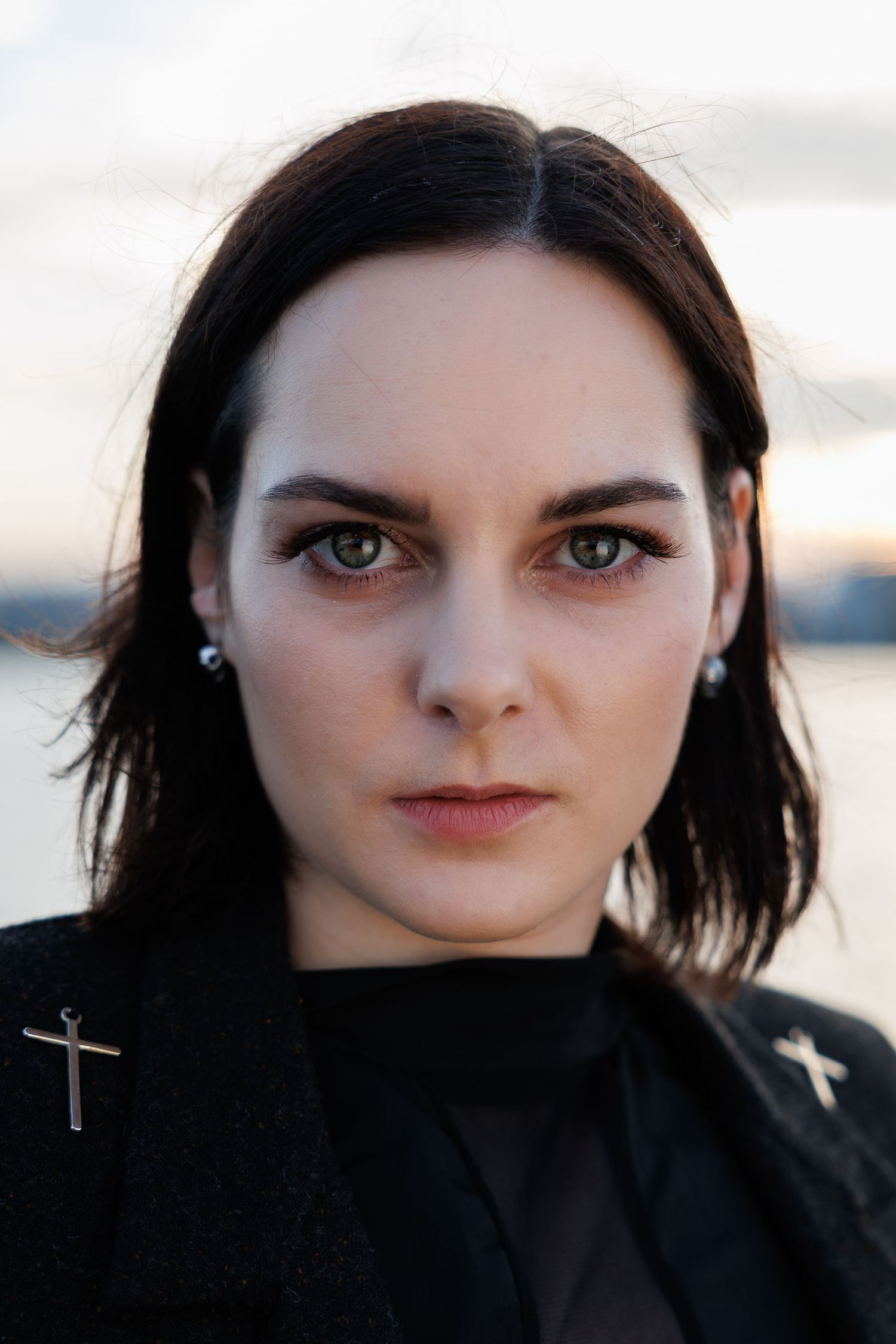 Young woman with dark hair and green eyes wearing a black jacket with cross pins near a waterfront at dusk.