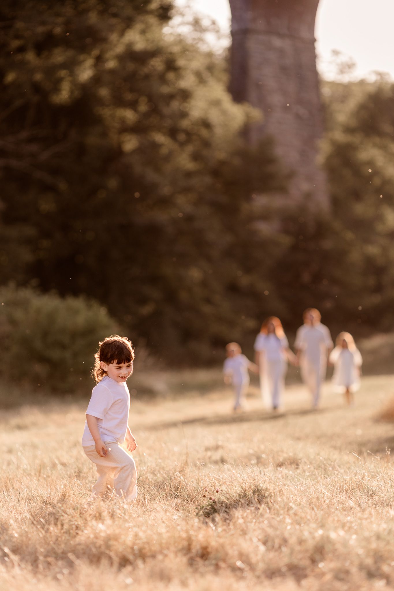 Children in white clothing play in a sunlit field with golden hour light and lush green trees in the background.
