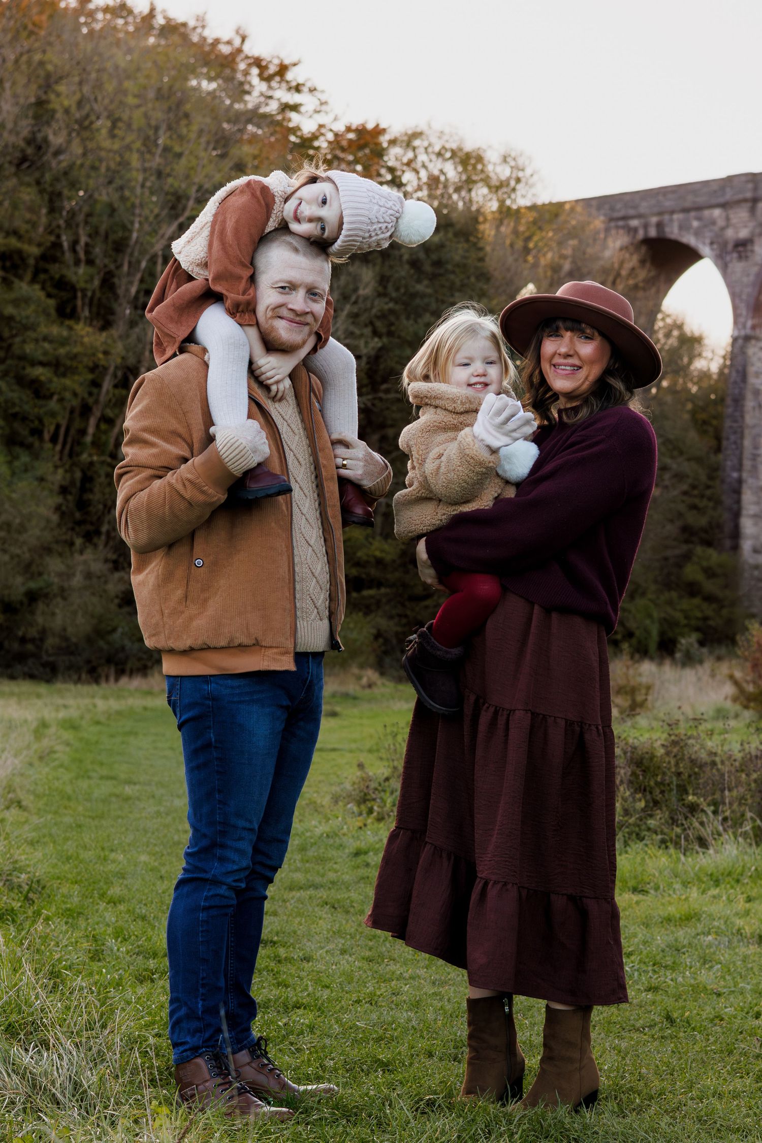 A happy family of four poses outdoors near a stone viaduct in autumn, wearing warm cozy clothing.