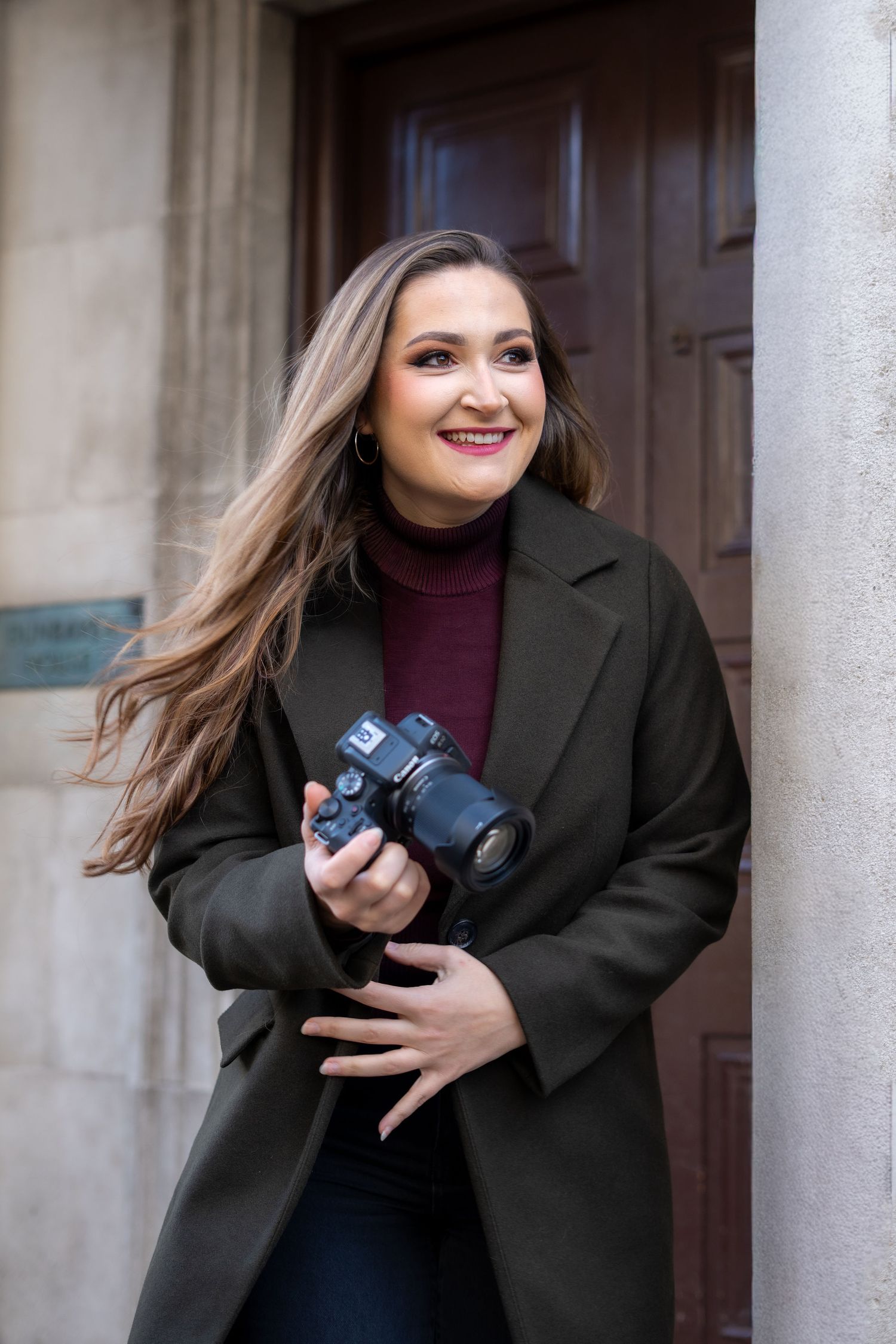 Smiling woman holding a DSLR camera while leaning against a stone building in an olive coat.