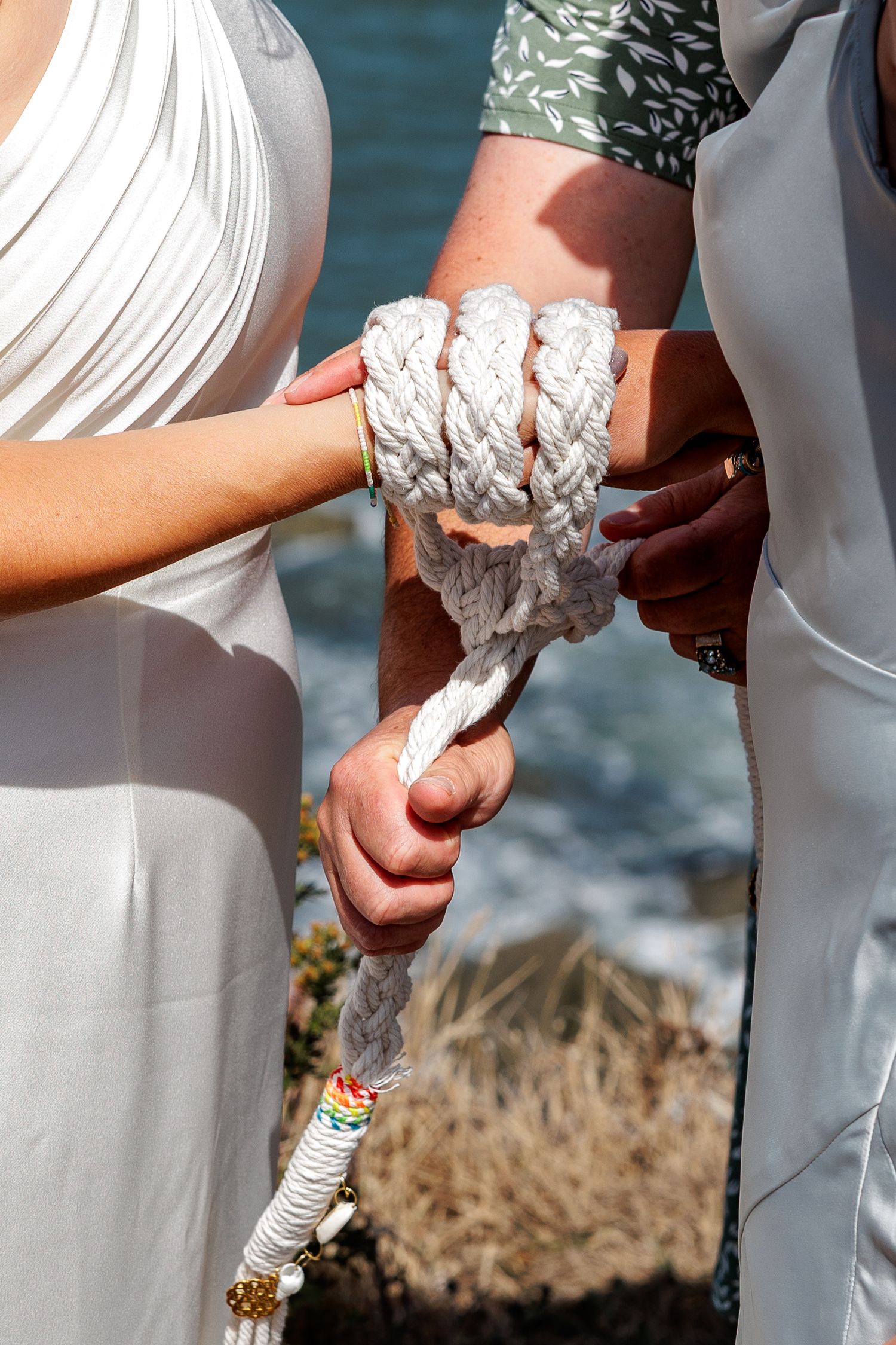 Handfasting ceremony with couple's wrists bound together by white rope near ocean cliffs.