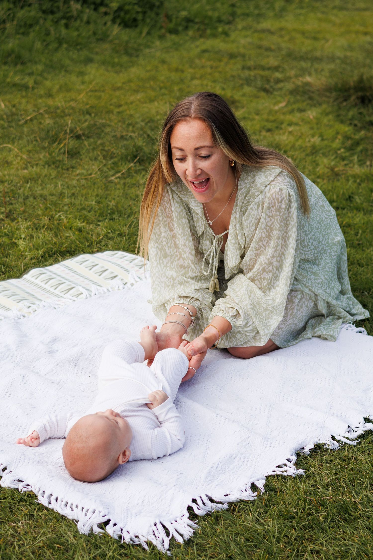 Mother playing with her baby on a white blanket in a lush green park, smiling joyfully outdoors.