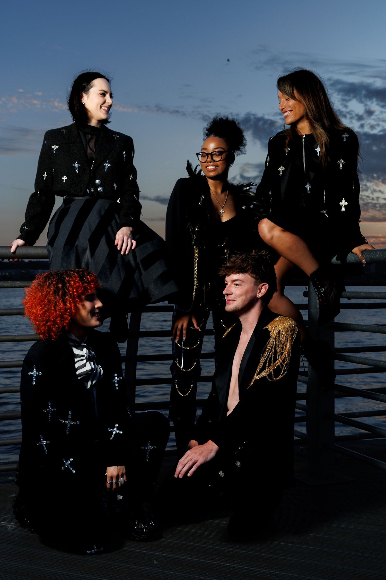 Five people in black cross-embellished fashion pose dramatically on a pier at dusk.