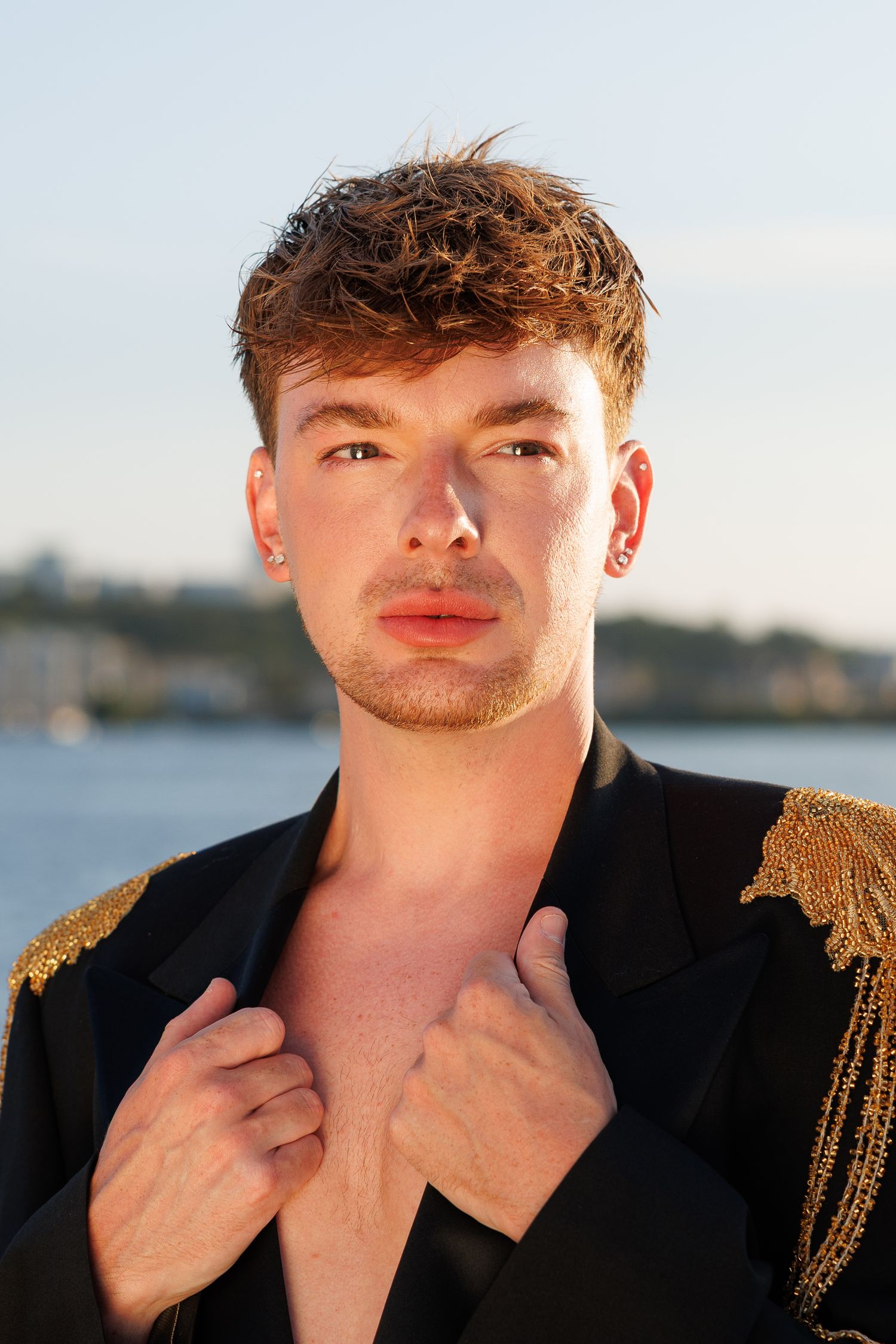 Young man in black gold-trimmed blazer poses outdoors near water at golden hour.