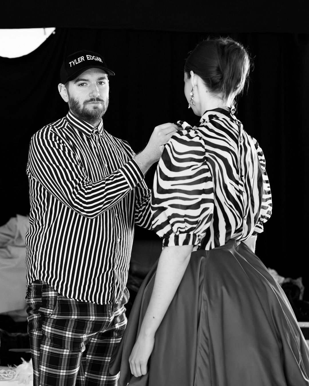 A fashion designer in a striped shirt adjusts a model's zebra-print top backstage at a fashion show.
