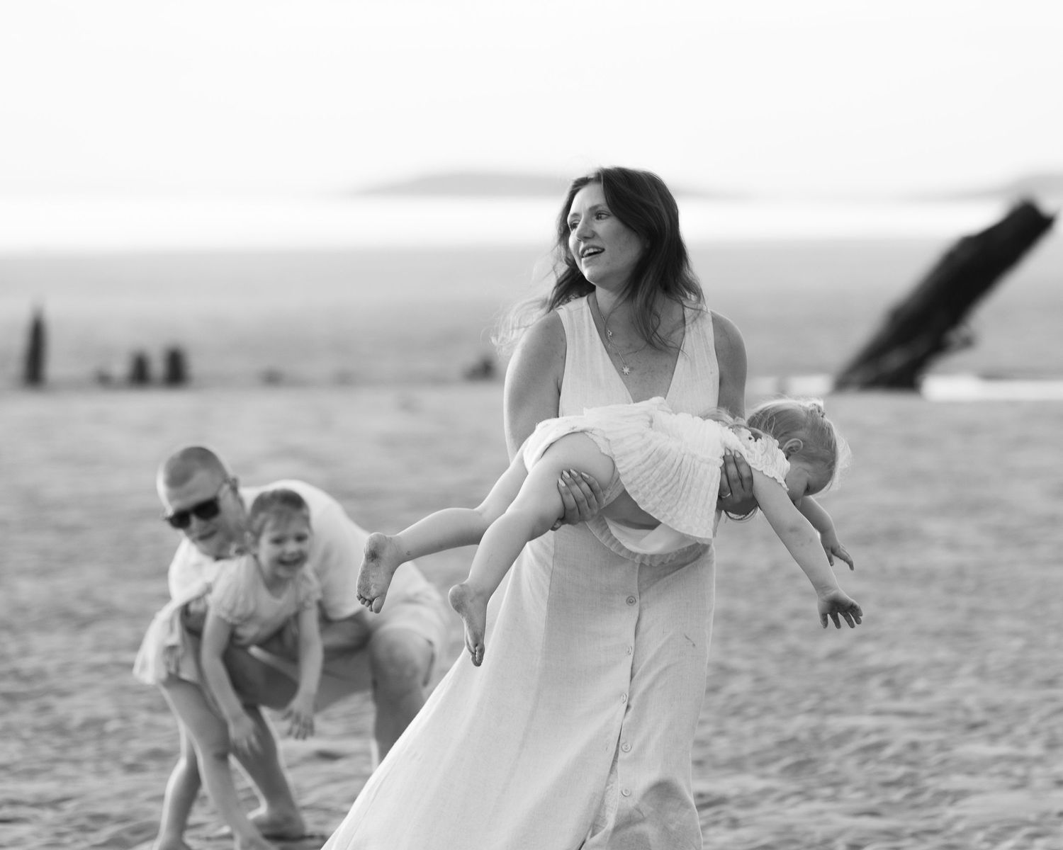 Black and white beach photo of a laughing woman in white dress carrying a child while others play behind her.