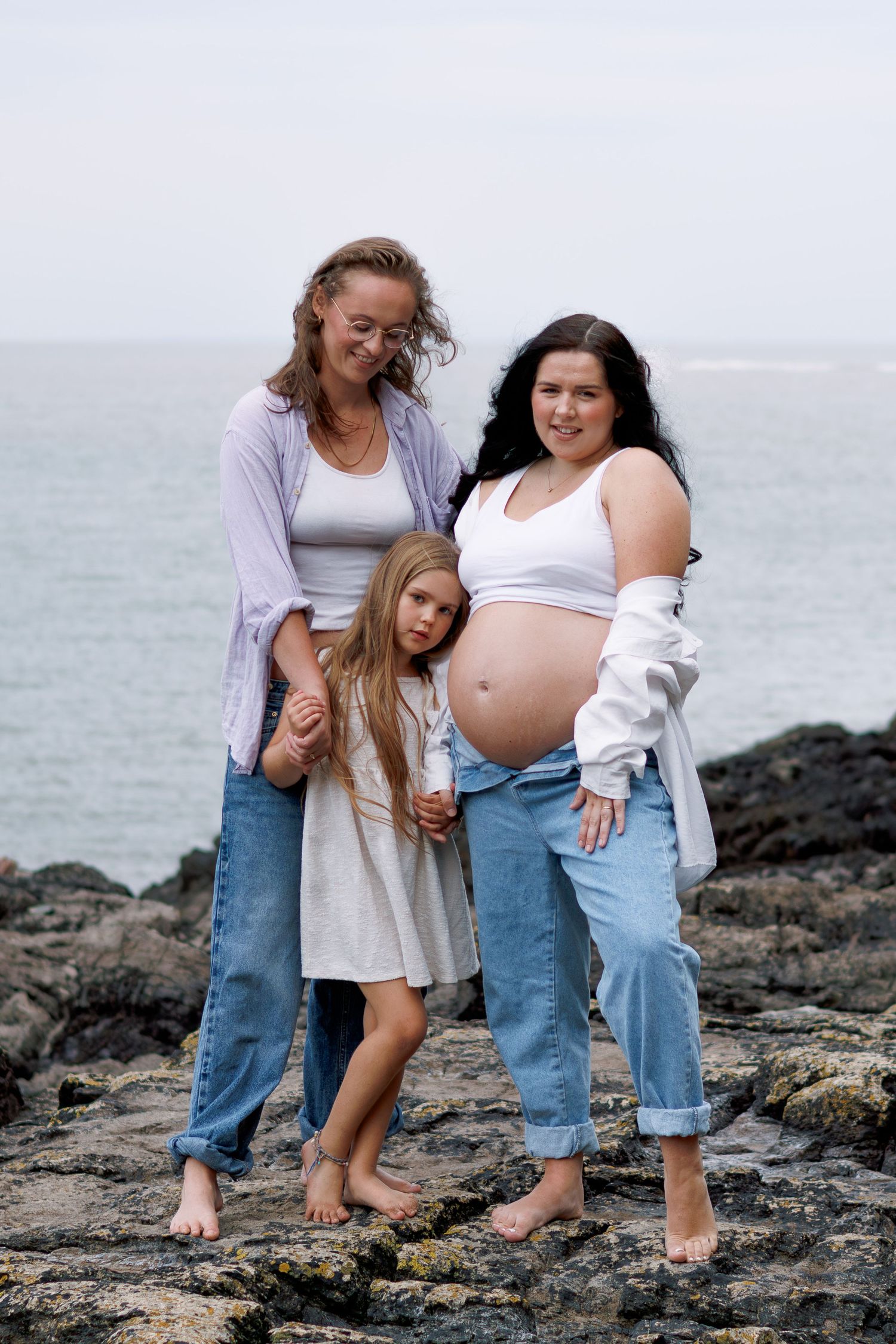 Two women and a young girl pose on rocky shore, one woman visibly pregnant, all wearing jeans and white tops.