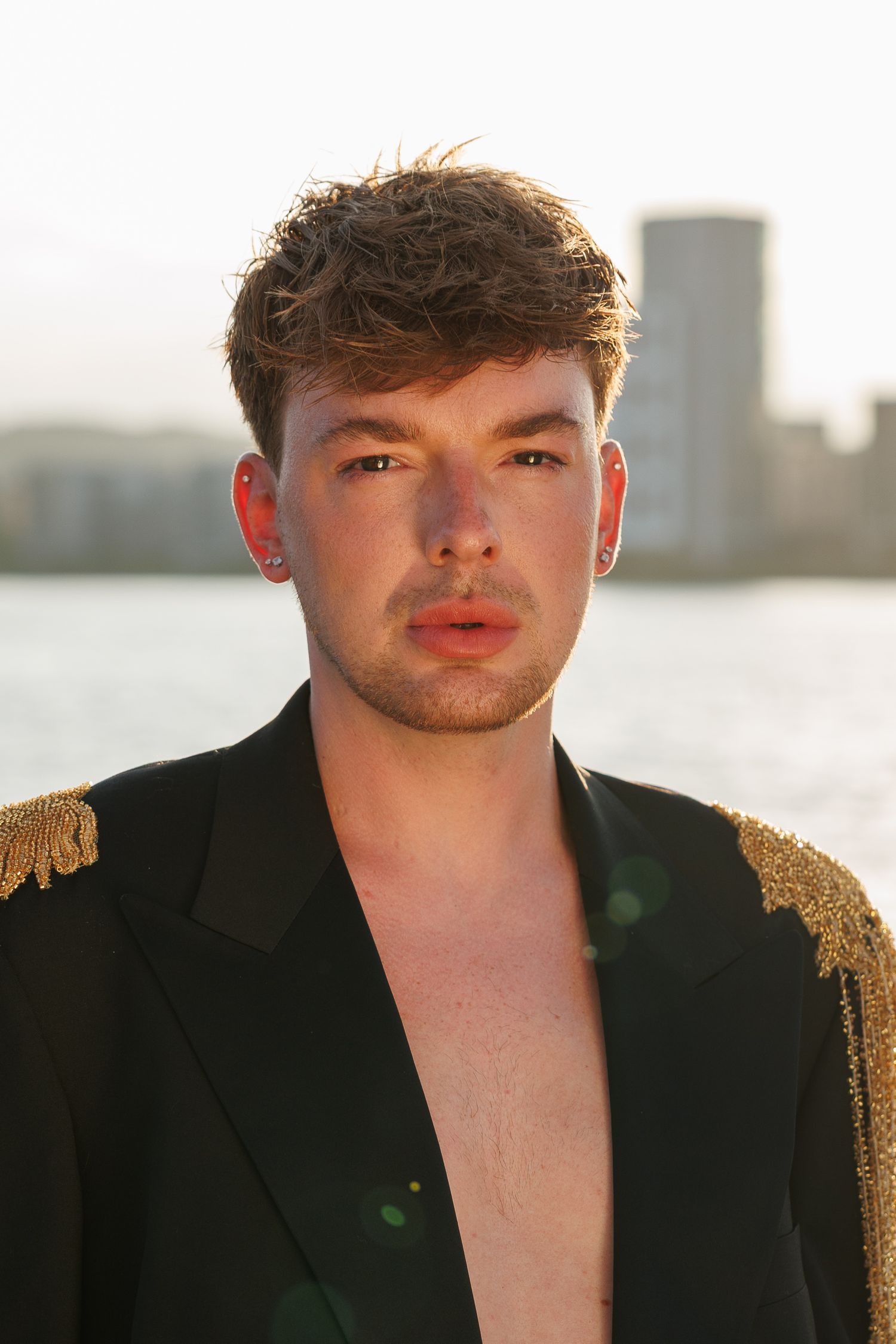 Young man in open black blazer with gold epaulettes poses outdoors near waterfront with city skyline backdrop.