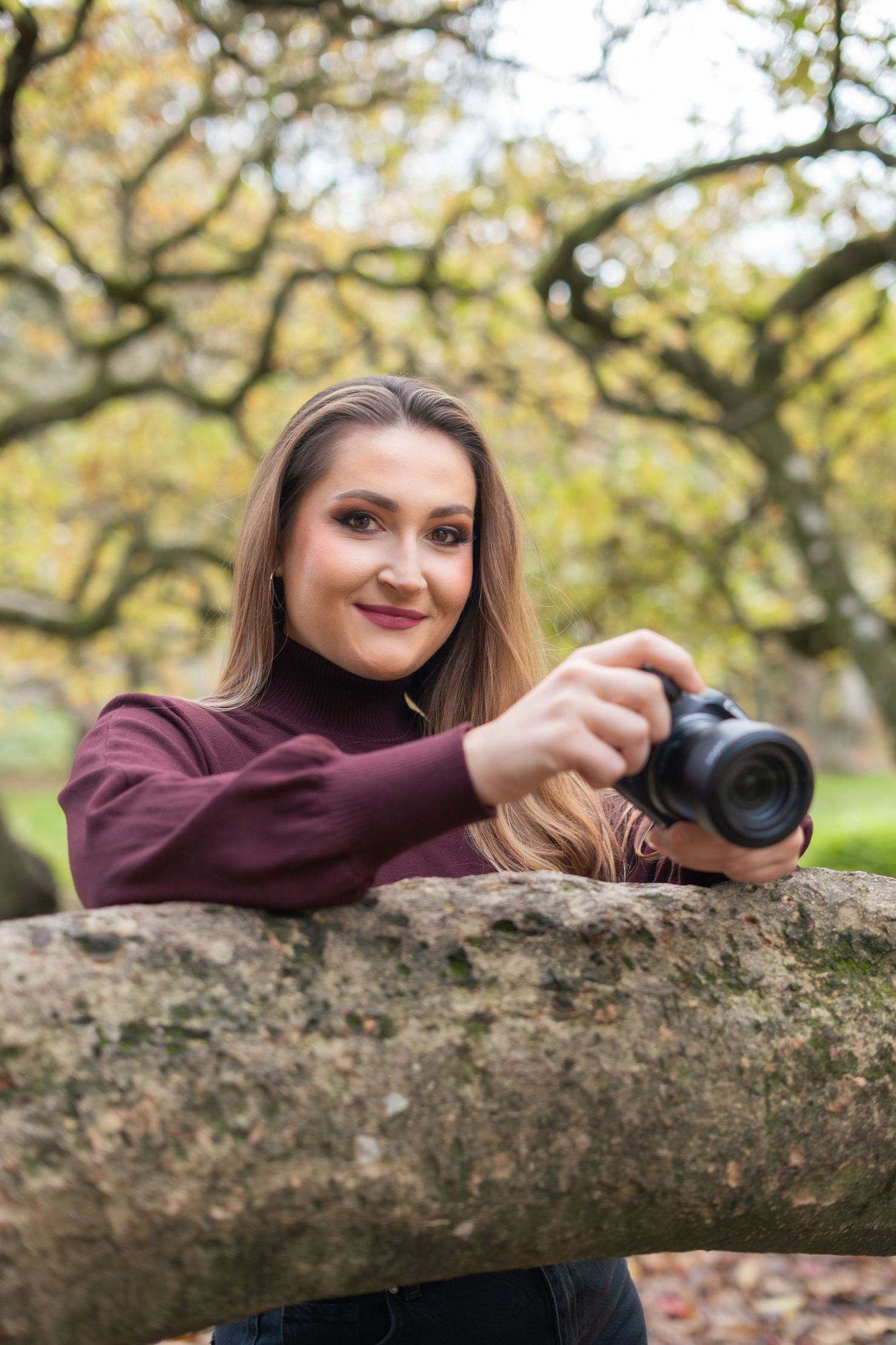A smiling woman in burgundy turtleneck holds a DSLR camera resting on a tree branch.