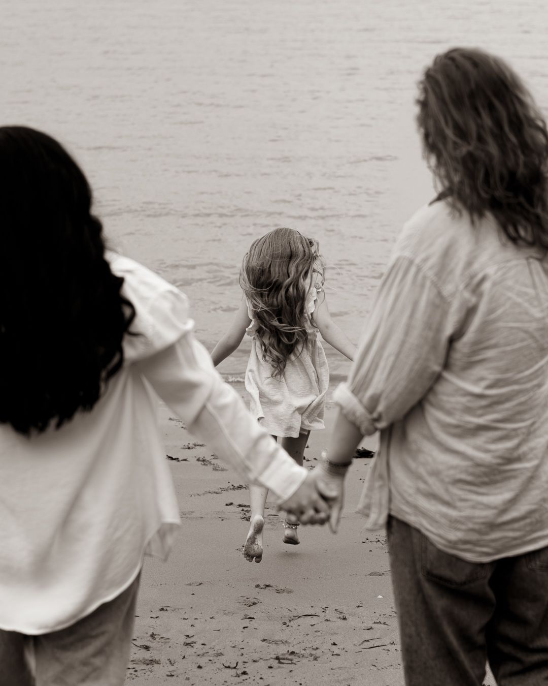 Two adults hold hands with a young girl on a beach in a black and white photo.