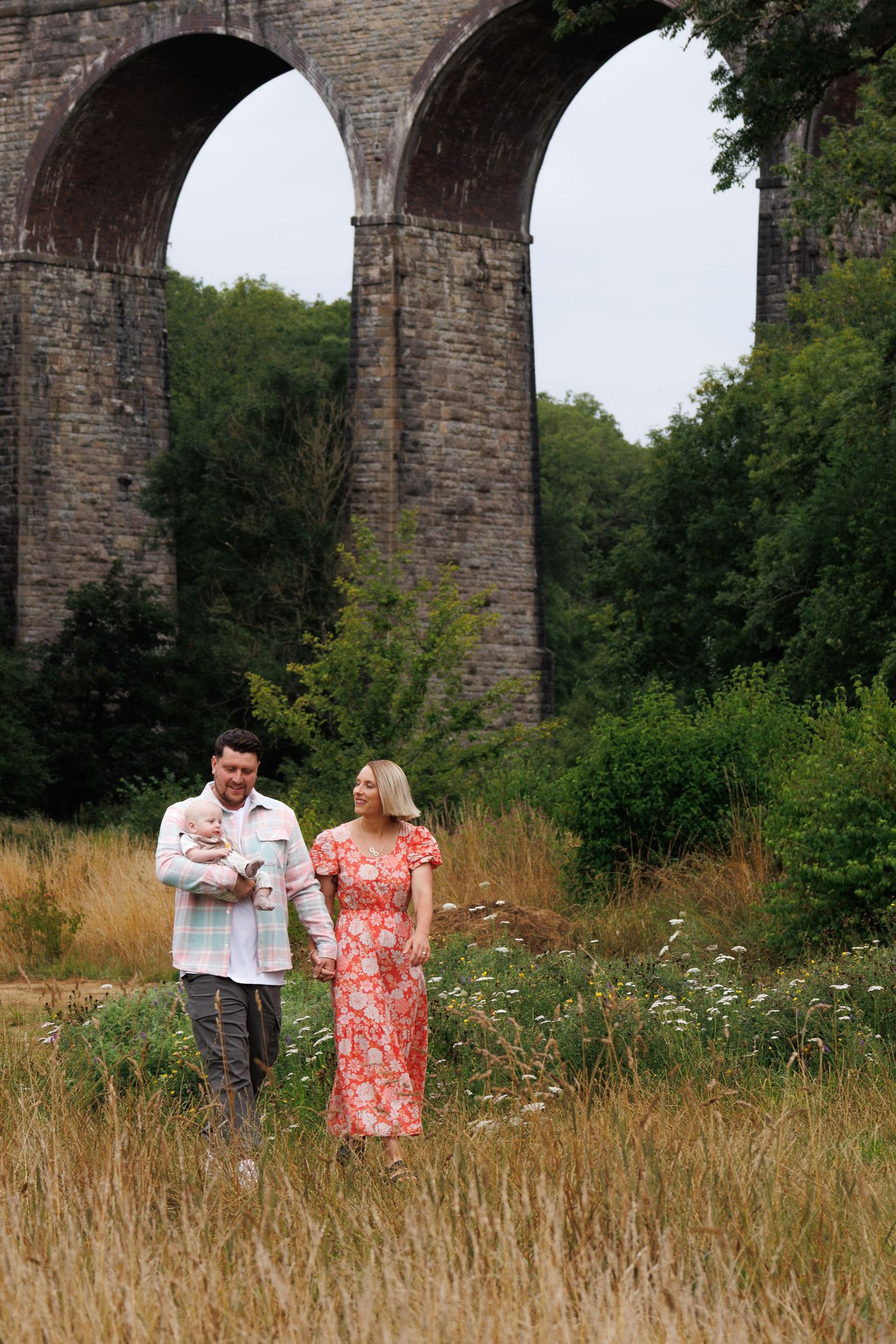Family with baby strolling in a meadow before a historic stone viaduct arch bridge surrounded by lush greenery.