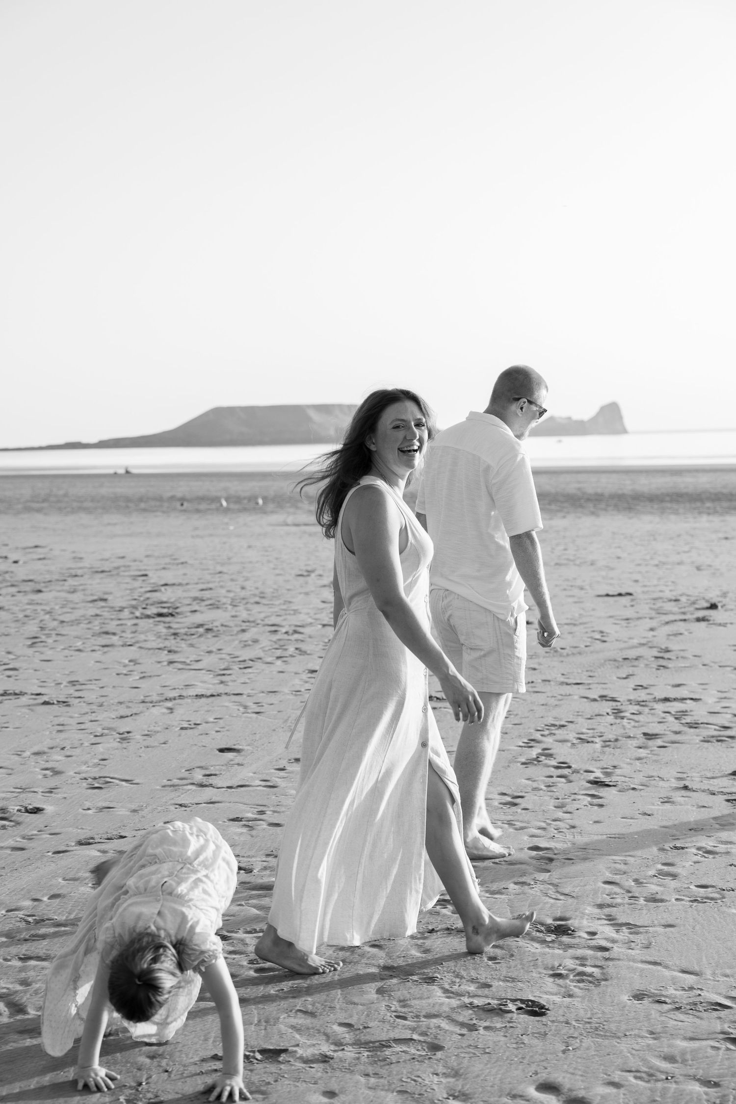 Black and white photo of a family in white outfits on a beach, woman smiling, man walking, child playing in sand.