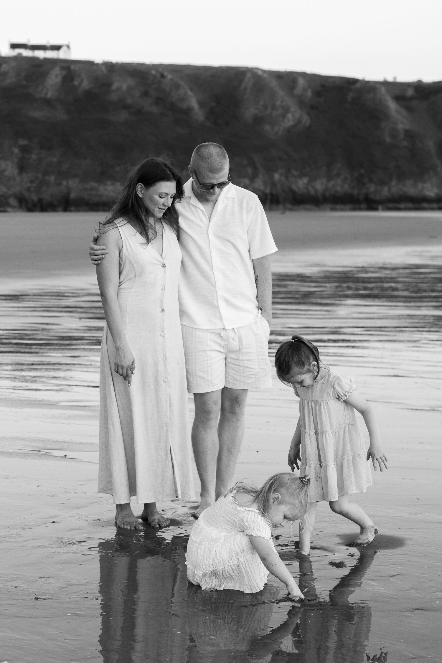 Black and white family beach photo with parents watching two young girls play in the water near rocky cliffs.