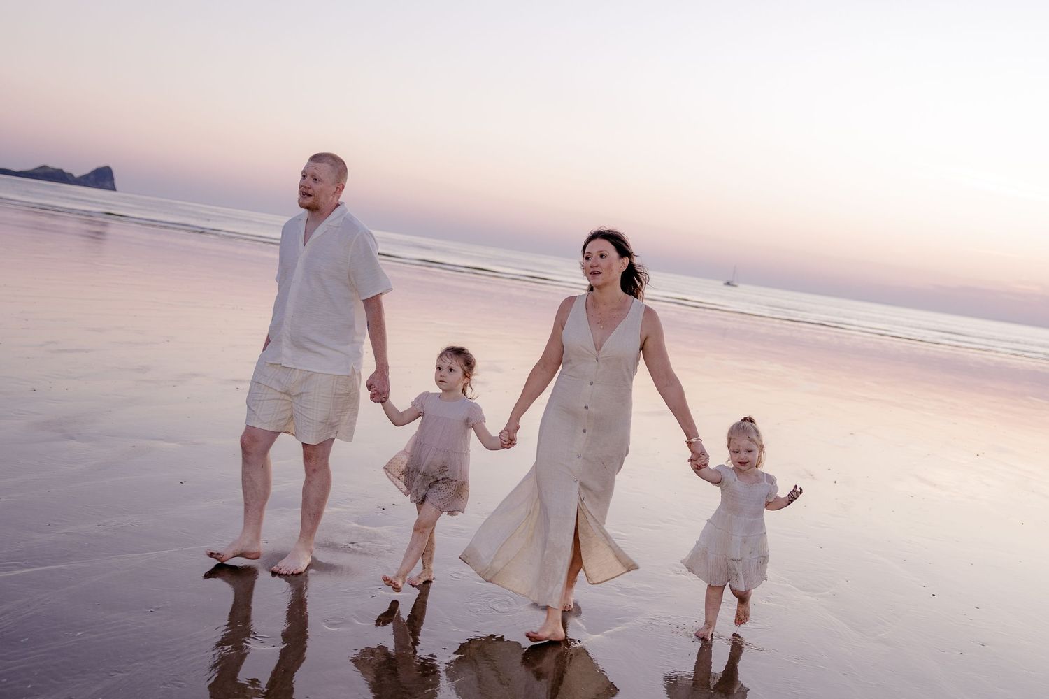 Family of four walking hand-in-hand on a beach at sunset, parents and two young girls in white outfits.