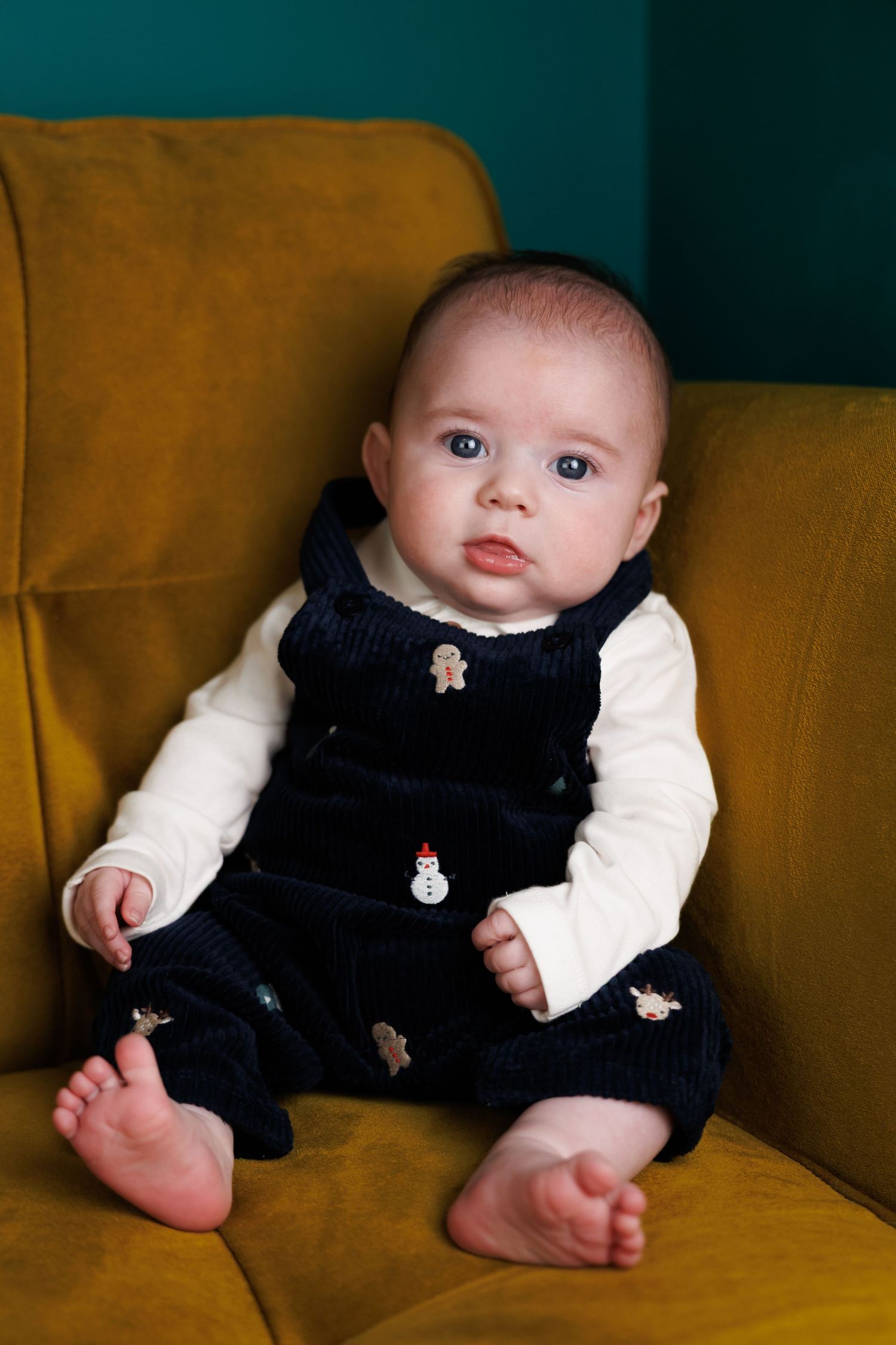 A cute baby in a navy Christmas jumper with festive embroidery sits on a mustard yellow armchair.