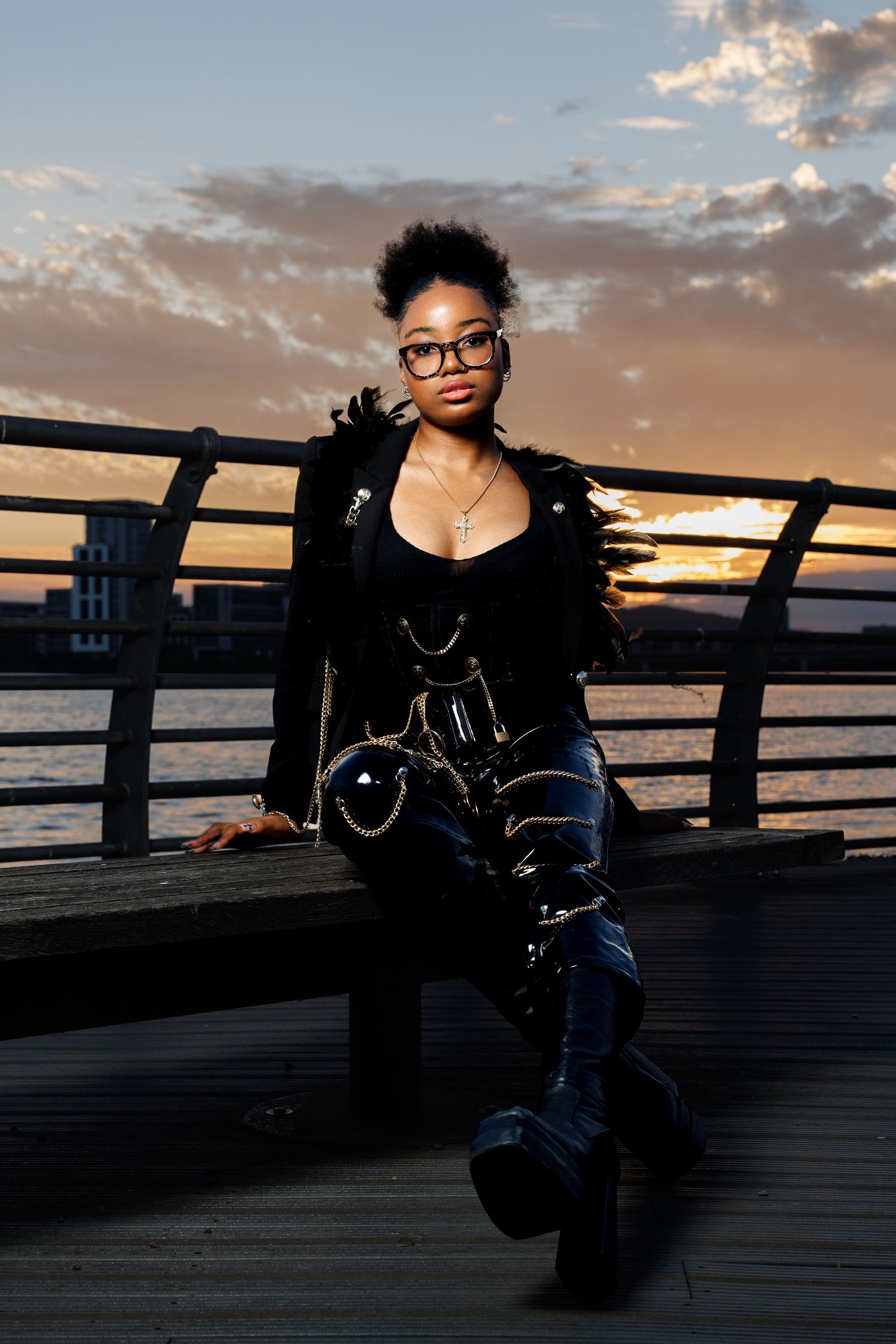 Young woman in black edgy fashion sits on pier bench at golden hour sunset.