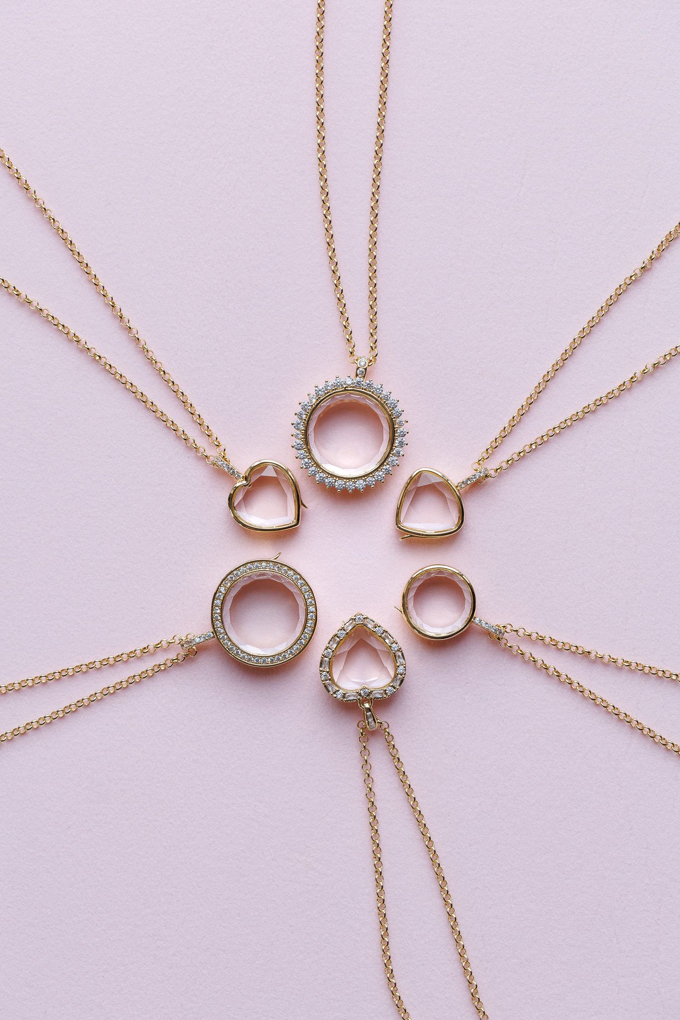 Five gold locket necklaces arranged in a sunburst pattern on a pink background, featuring circle and heart pendant designs.