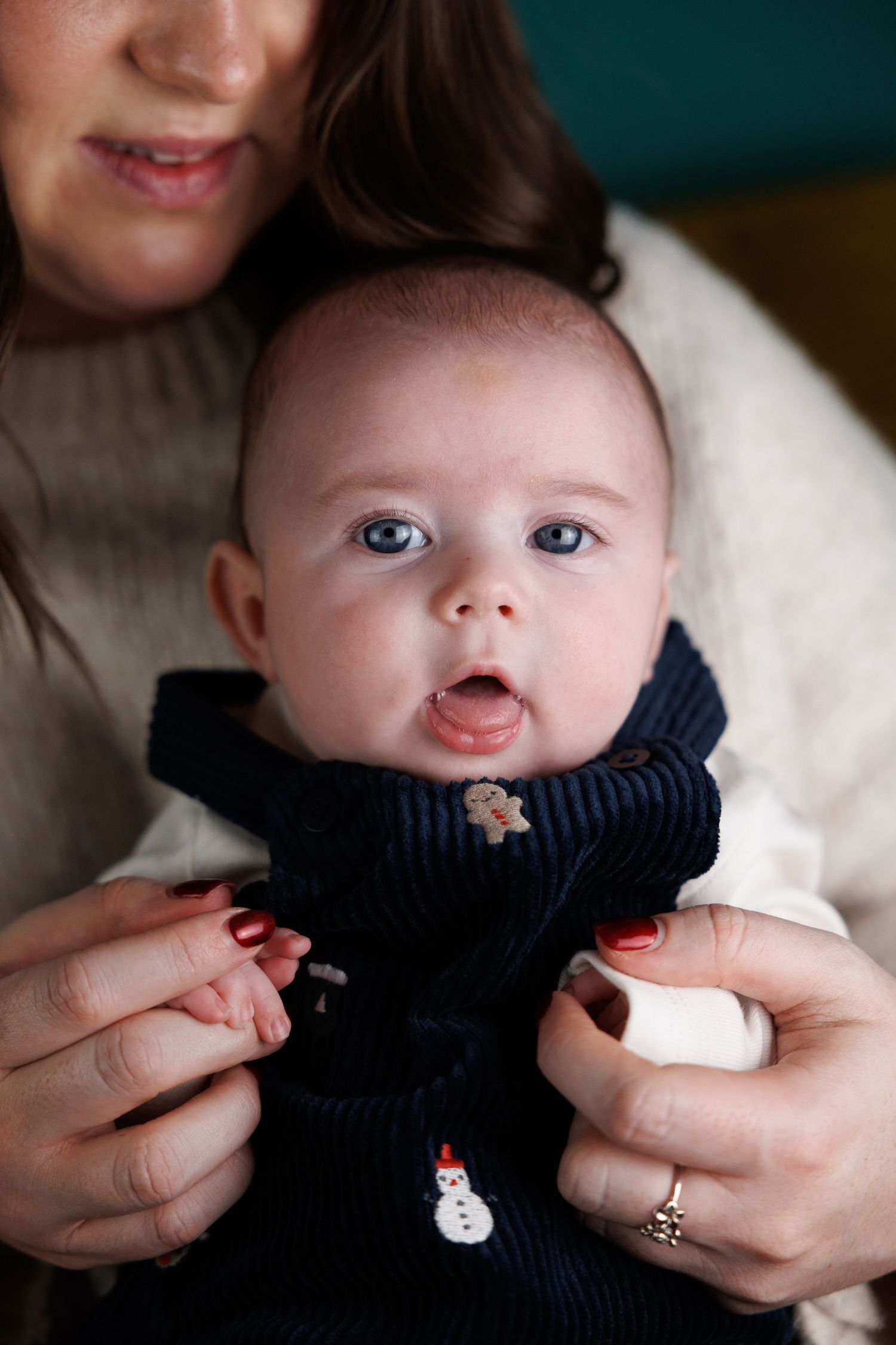 A wide-eyed baby in a navy Christmas sweater being held by a woman with red nails and a gold ring.