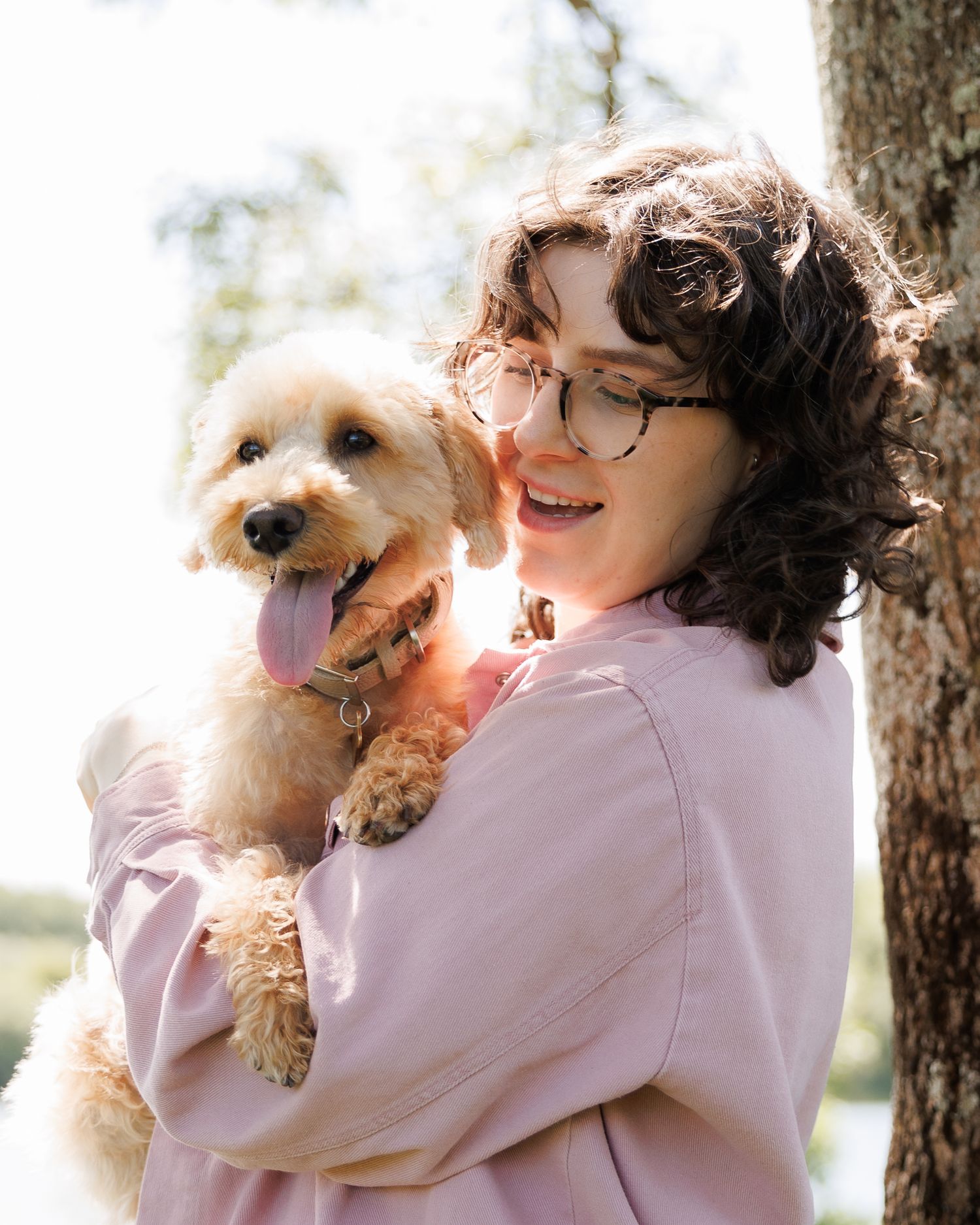 A smiling young woman with curly hair and glasses holds a fluffy golden doodle puppy outdoors near a tree.
