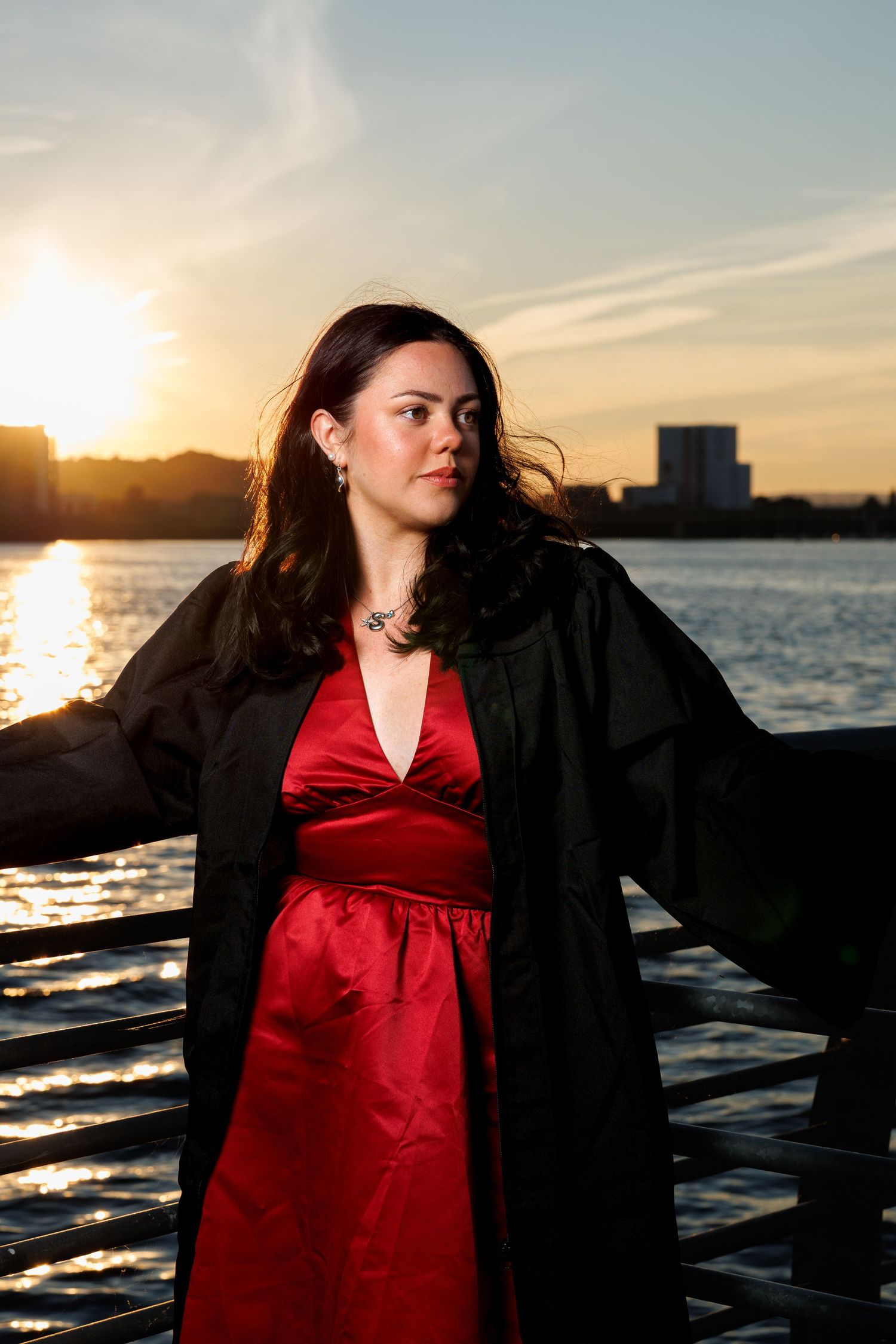 Woman in red satin dress and black graduation gown poses by waterfront at sunset with city skyline.
