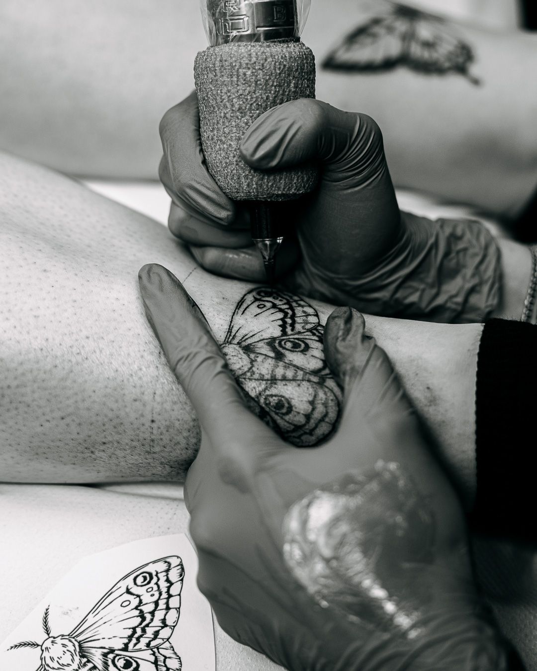 Close-up black and white photo of tattoo artist inking a butterfly design on skin.