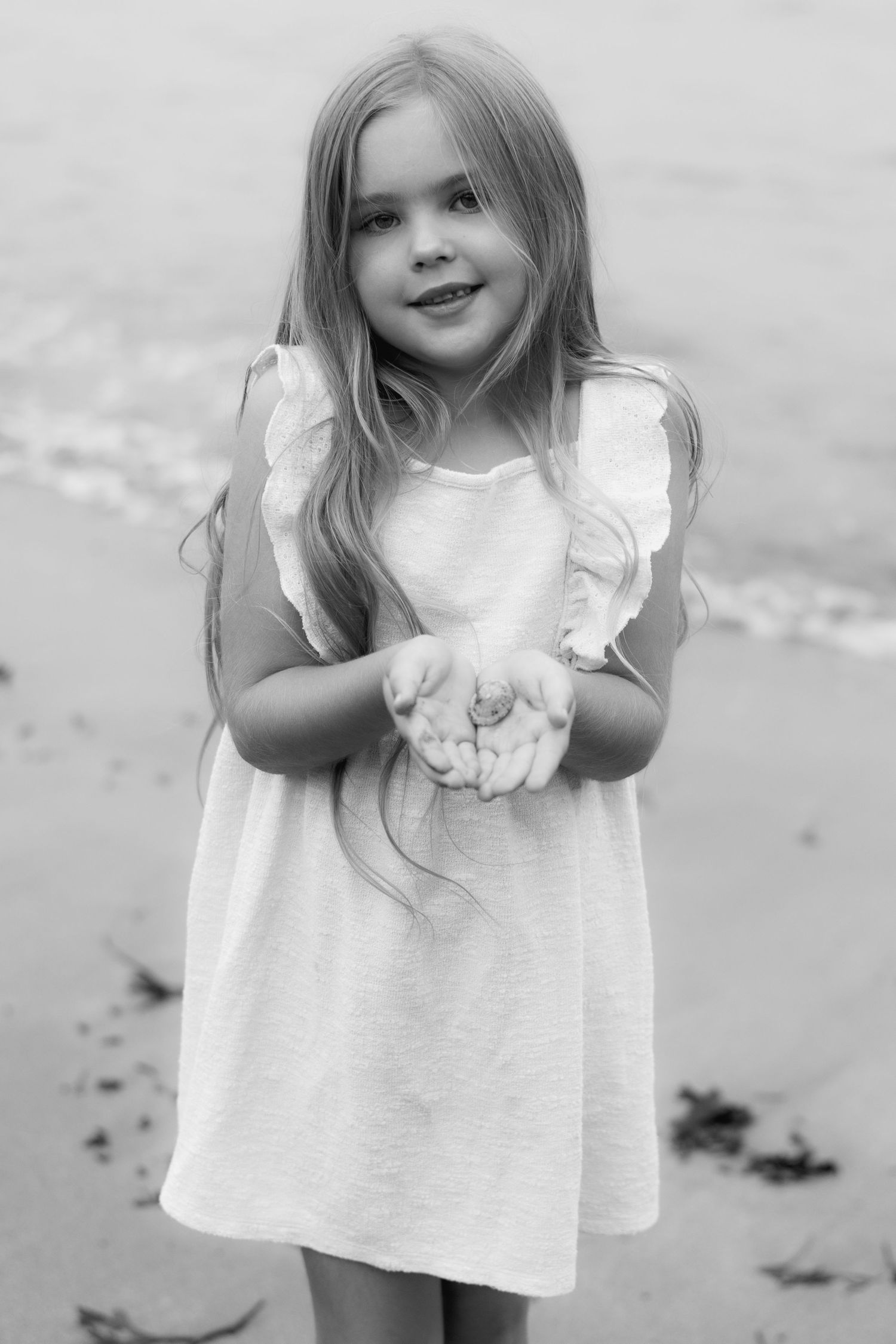 Black and white photo of a young girl in a white dress holding a seashell on a beach, smiling at the camera.