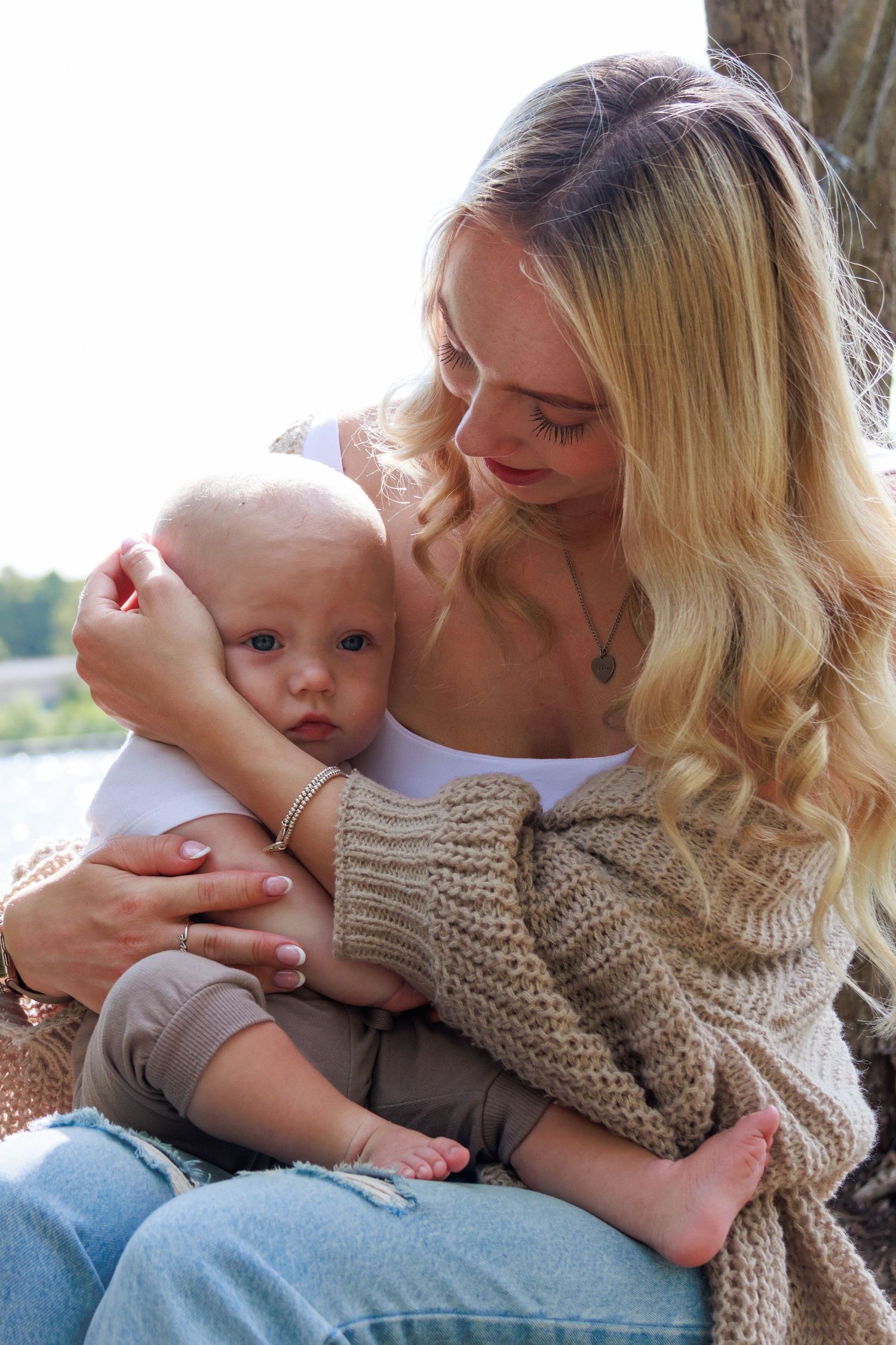 Blonde mother tenderly holding her baby infant outdoors near water, wearing a cozy knit cardigan.