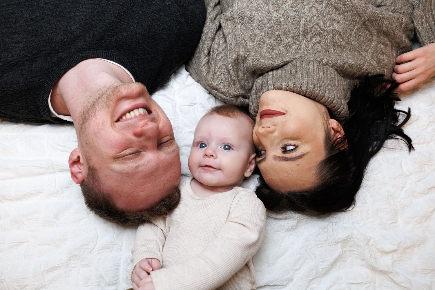 Happy family portrait with parents and baby lying on white blanket, smiling together.