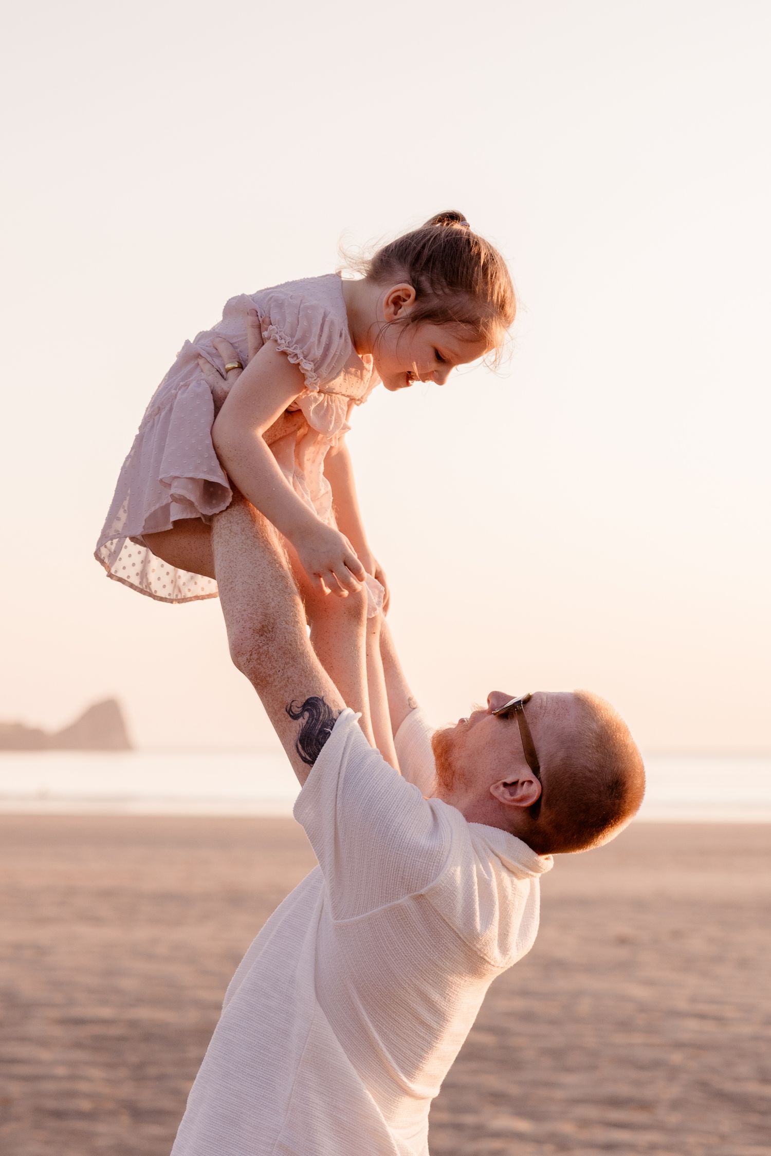 Father lifting young daughter in the air on a sandy beach at golden hour sunset.