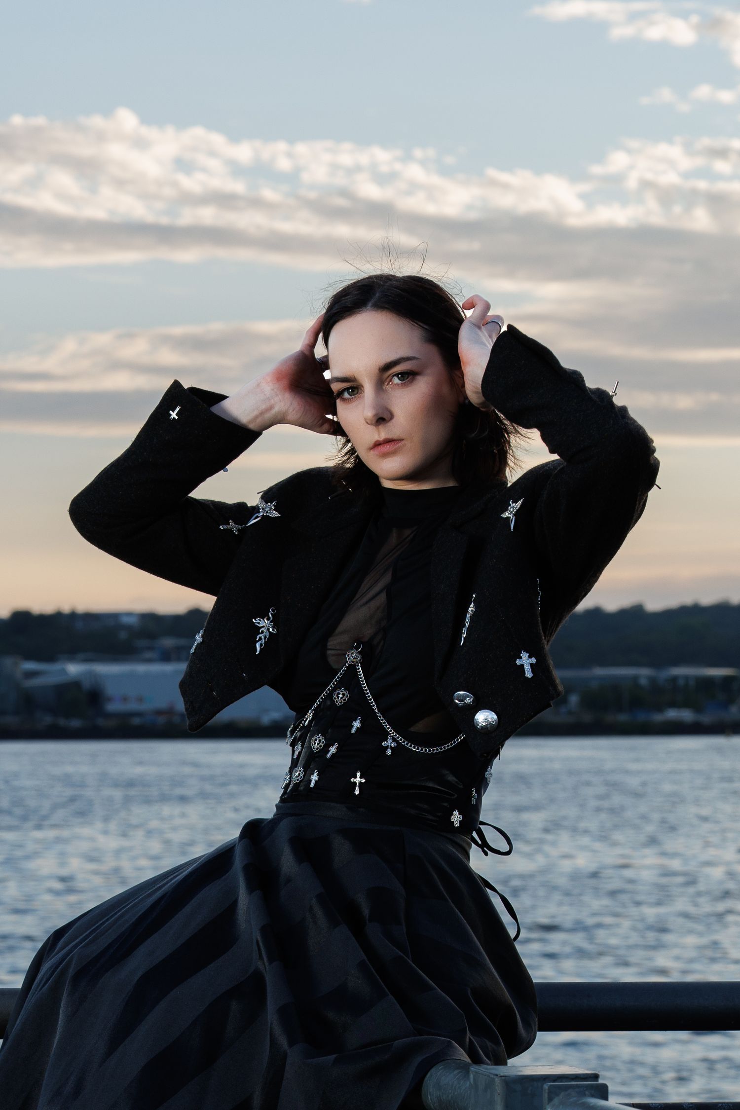 Young woman in gothic black outfit with cross embellishments poses by waterfront at dusk.