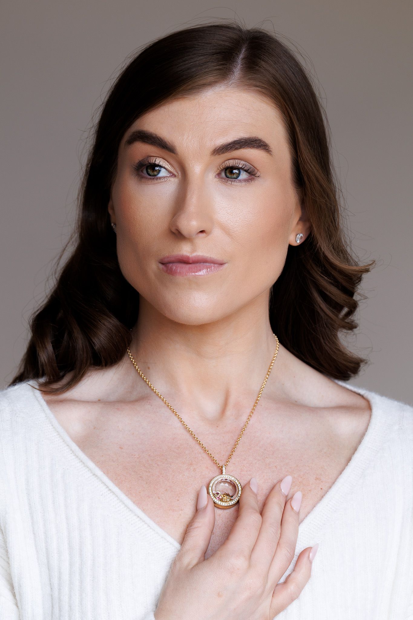 Woman wearing a gold circle pendant necklace and stud earrings, dressed in white, with dark hair and natural makeup.