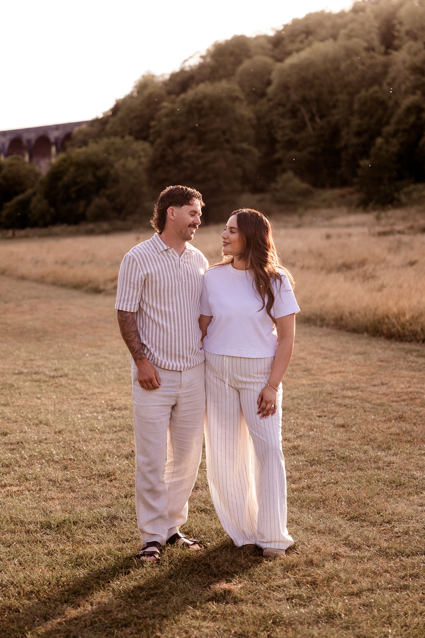A smiling couple in matching white striped outfits stand together in a sunlit field with green trees in the background.