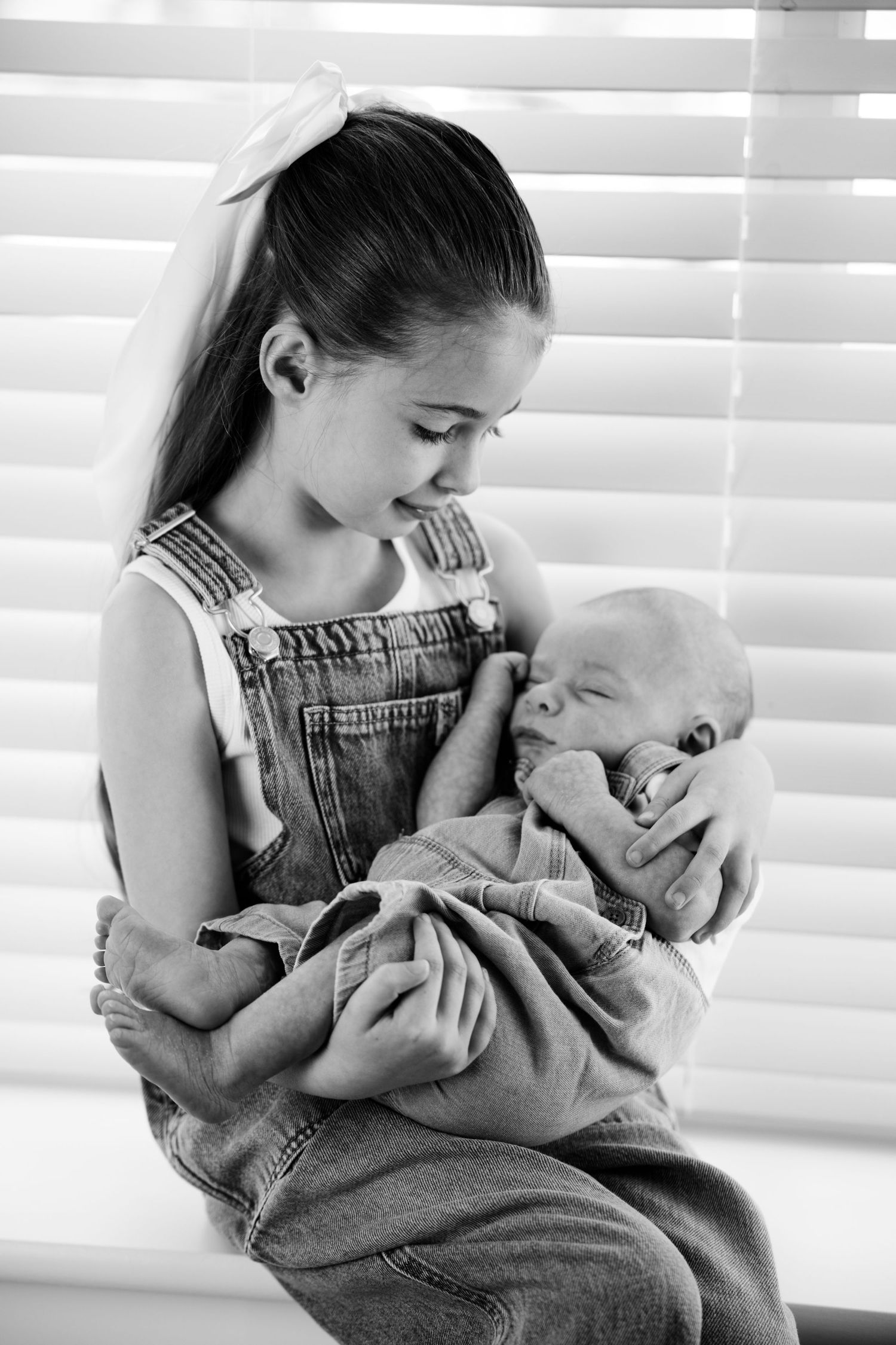 Black and white photo of a young girl in denim overalls tenderly holding a sleeping newborn baby near a window.