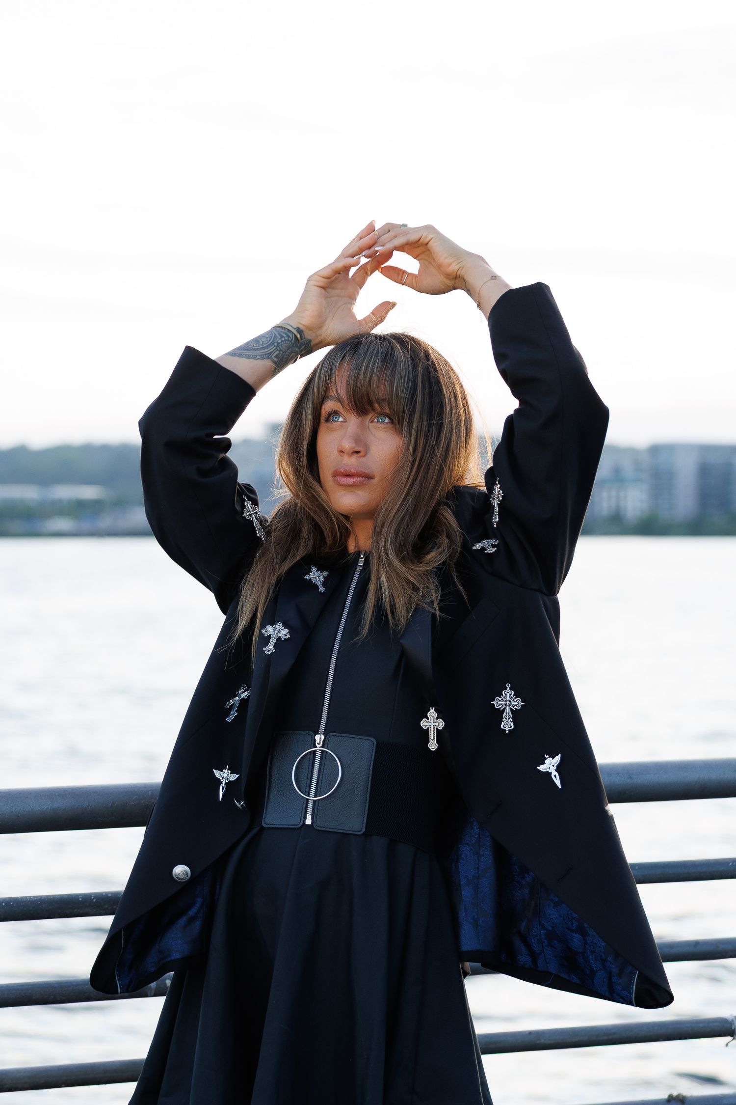 Woman in black cross-embroidered coat poses with arms raised near waterfront, wearing gothic fashion accessories.