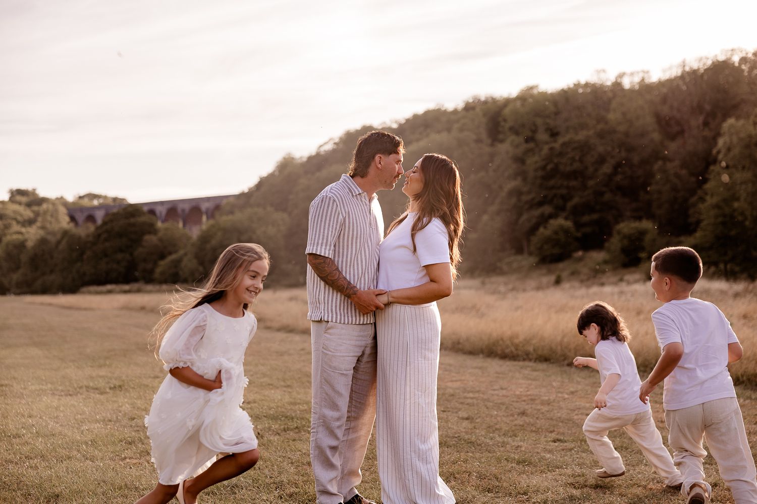 A pregnant couple shares a tender moment in a golden field while their children play around them at sunset.