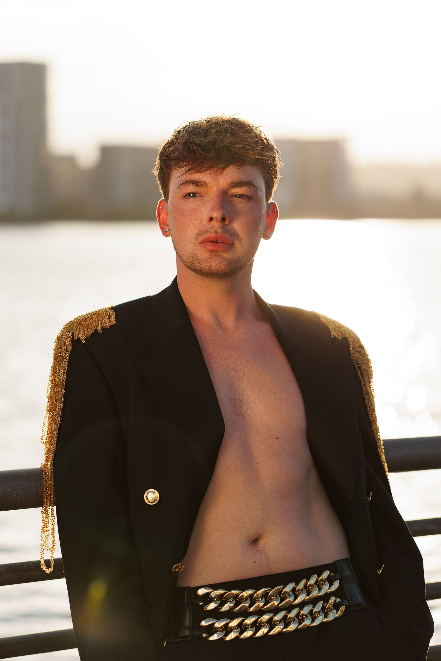 Young man in open black military jacket with gold epaulettes and chain belt posing by waterfront at golden hour.