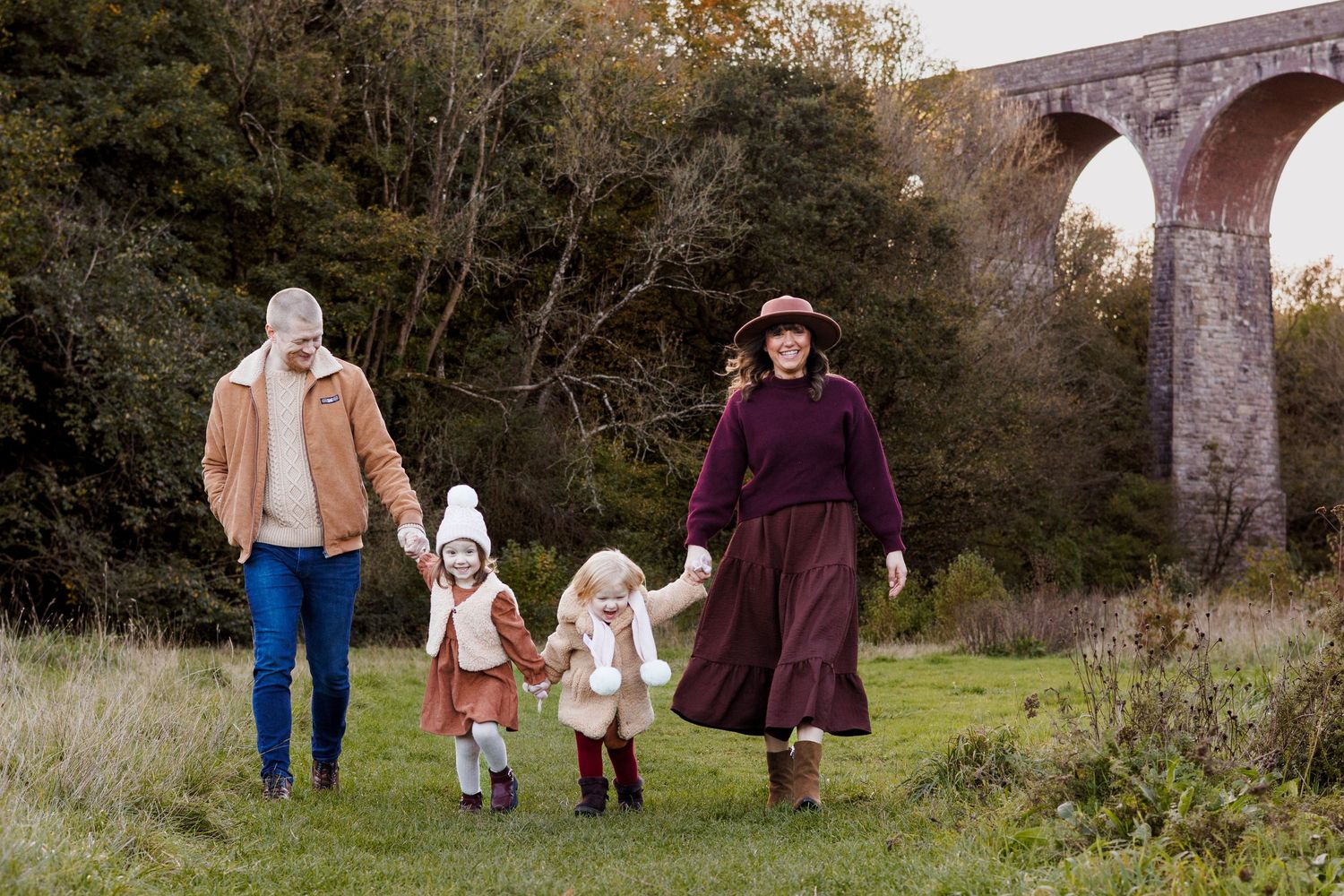 Family of four walking hand-in-hand through a green meadow with a stone viaduct and autumn trees in the background.