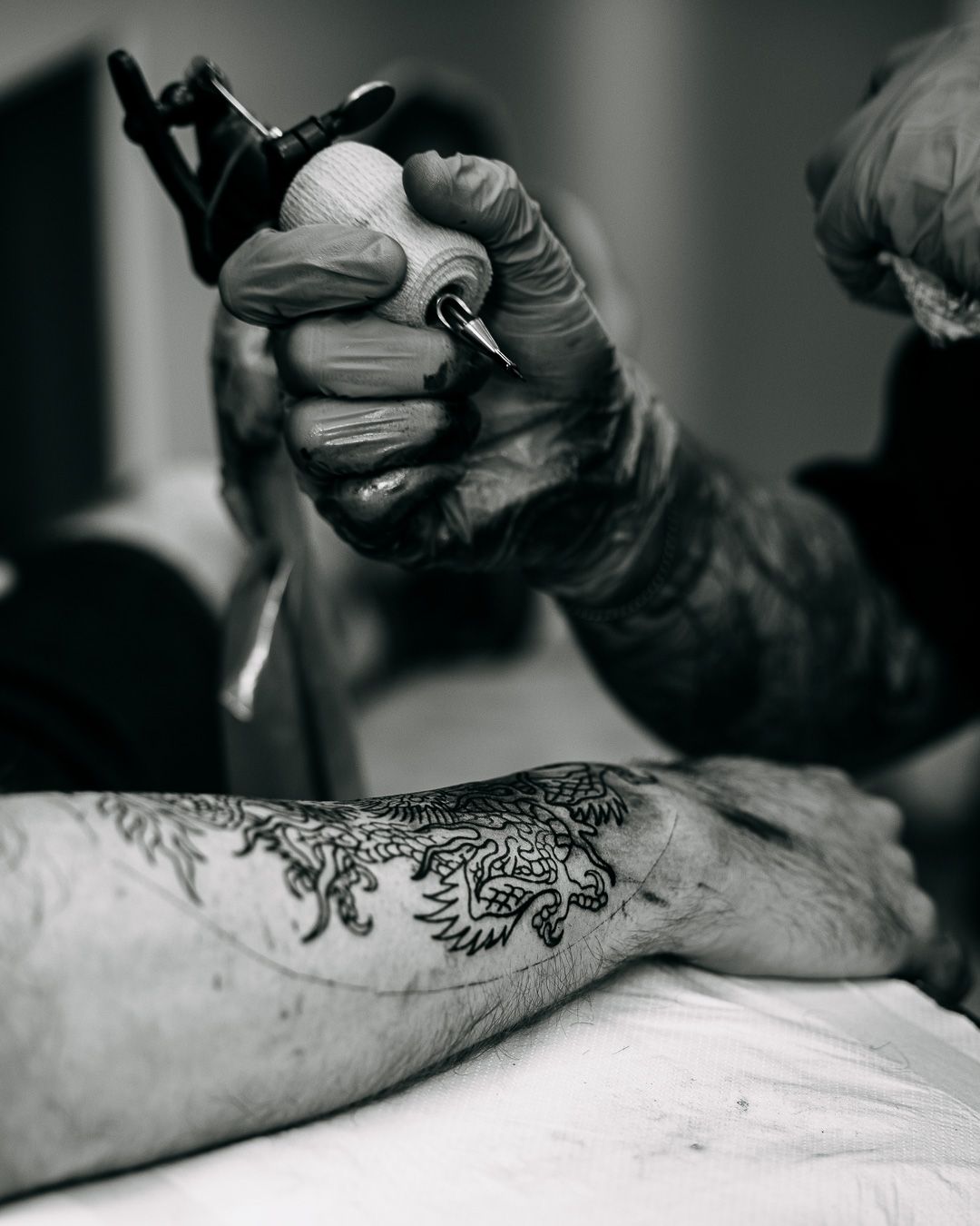 Black and white close-up of tattoo artist using machine to ink a lion design on forearm.