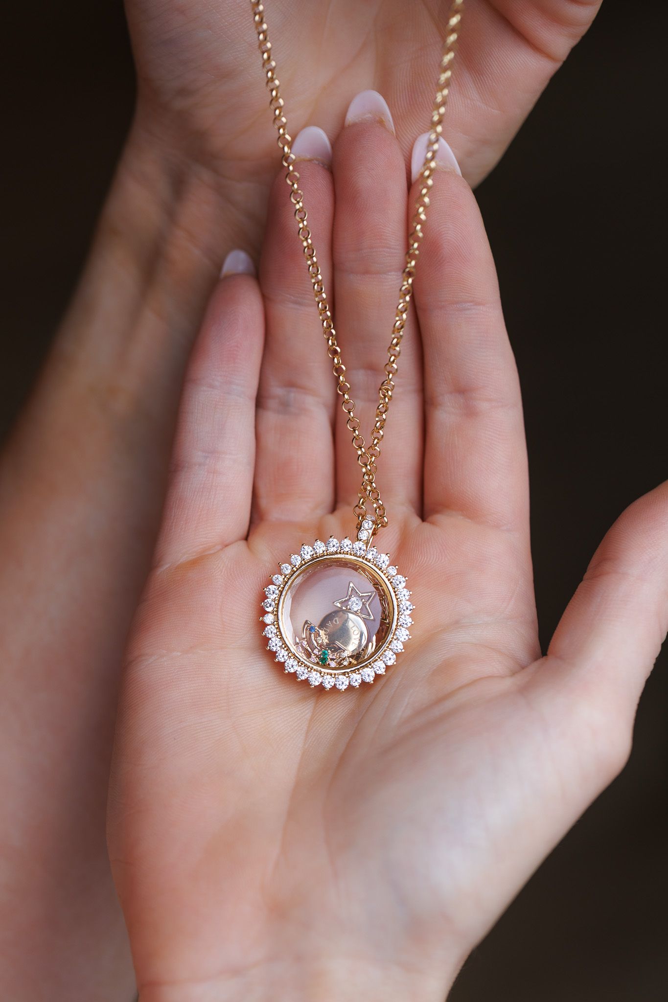 Hands holding a gold locket necklace with crystal halo pendant on delicate chain.