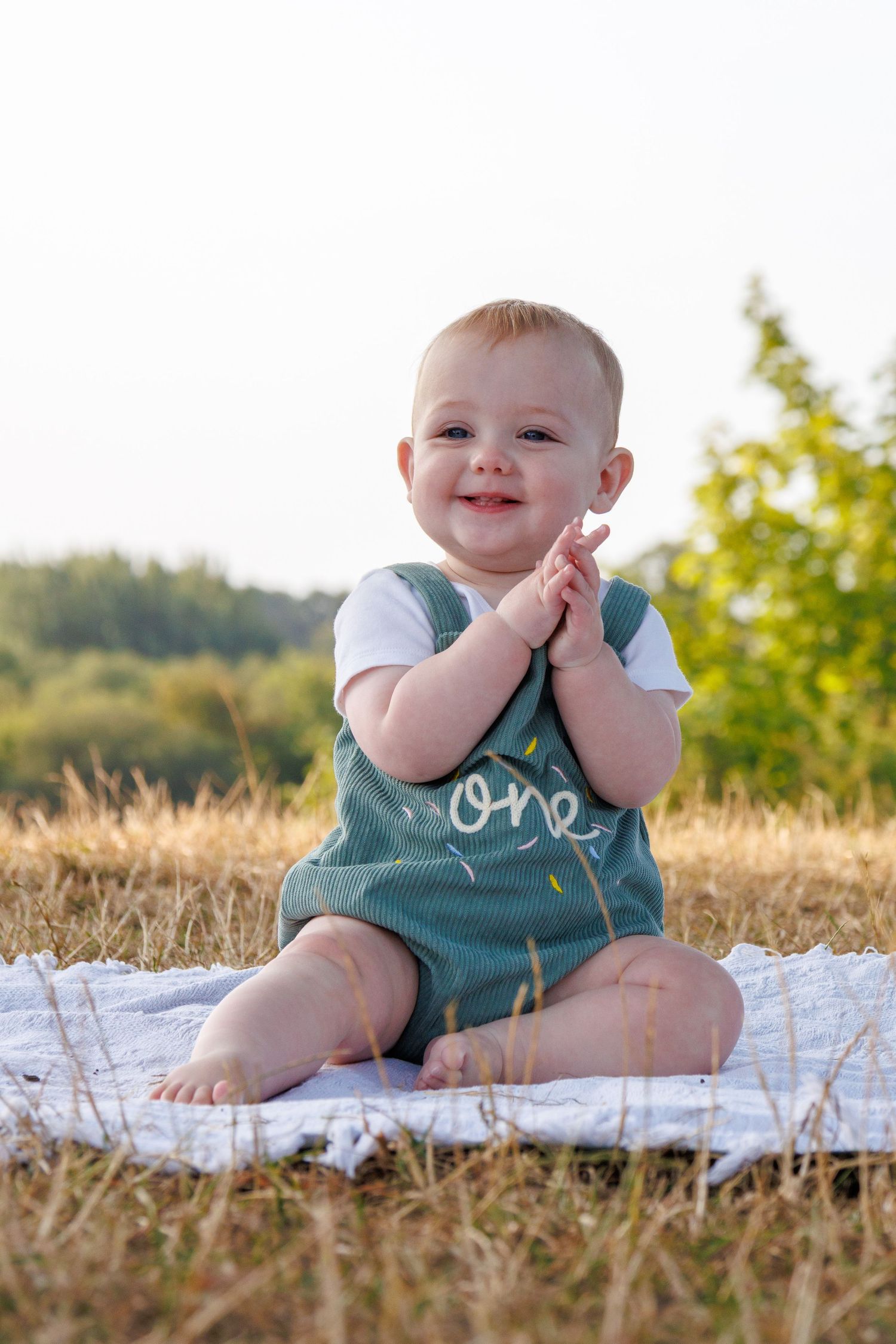 Happy baby in green overalls sitting on blanket outdoors, clapping hands and smiling on a sunny day.