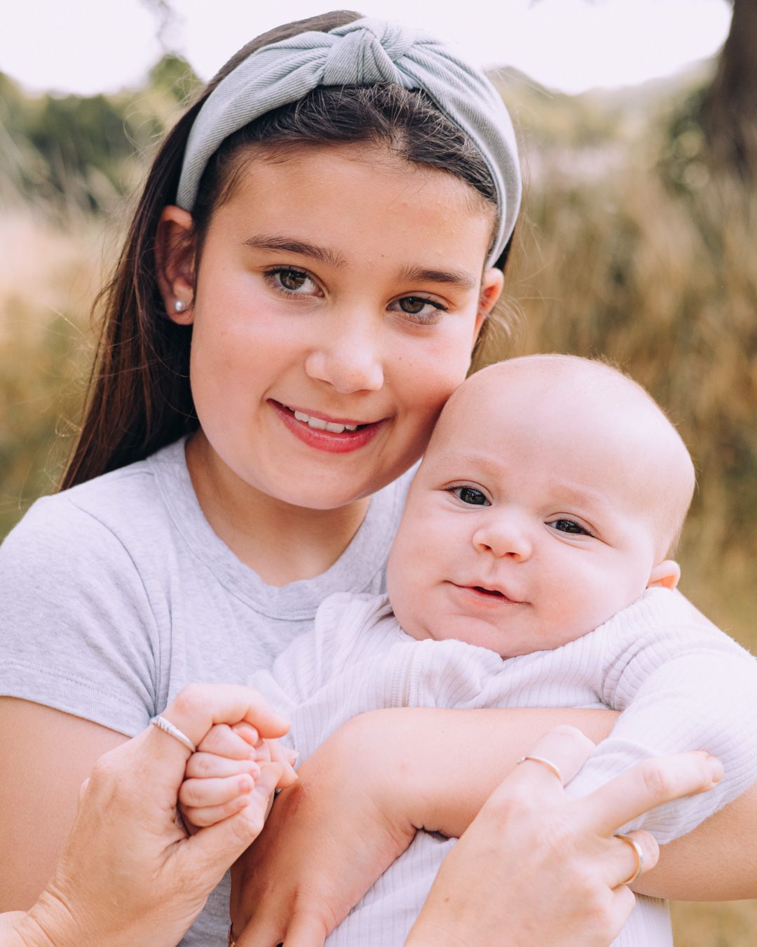 A smiling young girl with a gray headband holds an adorable baby in a white onesie outdoors.