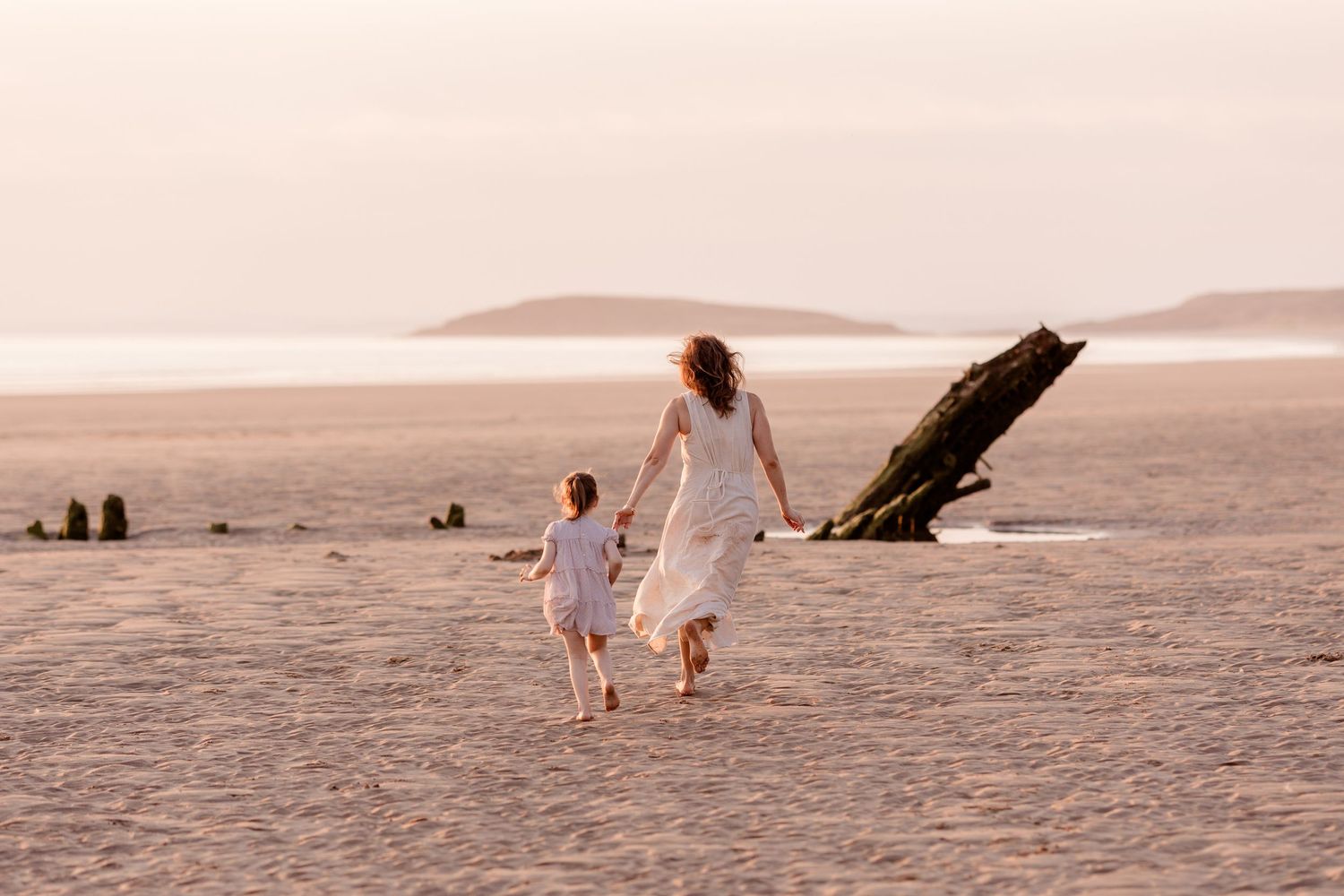 Mother and daughter in white dresses hold hands running on a sandy beach at sunset.